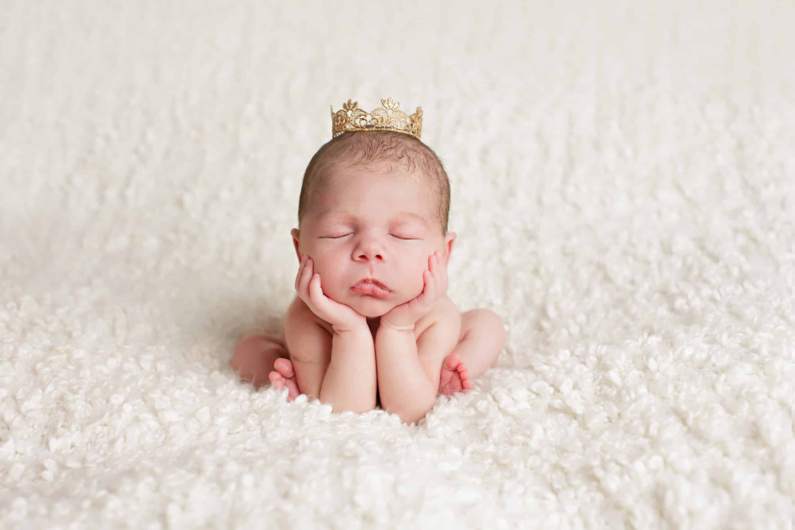 A newborn baby sits on a white blanket held up by its hands and elbows while wearing a gold crown