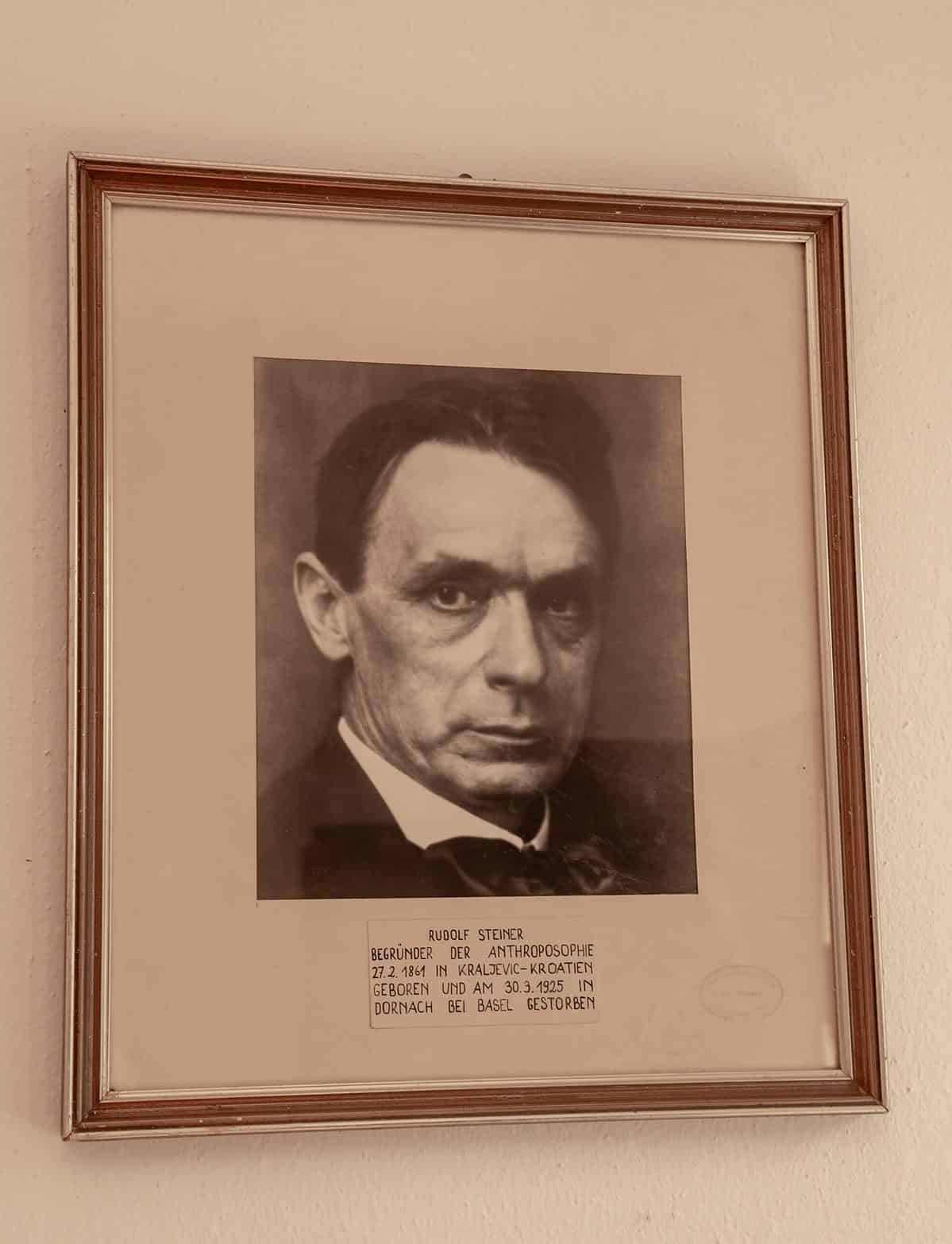 A photo of Rudolf Steiner in a frame with a mat. The frame is slender and possibly wood or plastic with gilt (gold) around its edges.  The mat is quite wide and below the photograph,  type-written on a piece of paper that matches the neutral tone of the mat has Steiner’s name and then some German words that are not distinct. The frame is hanging on a neutral wall. 