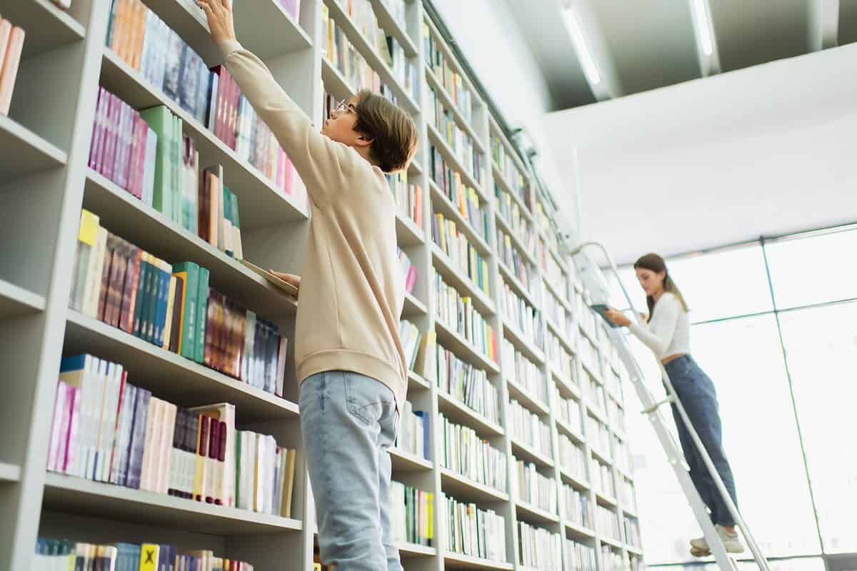 A low angle photograph of light gray wooden bookshelves filled with what appear to be textbooks. There are at least nine levels of shelves. In the back of the photograph toward the right is a young light-skinned woman with long brown hair, wearing a long sleeve white crop top and high waisted denim jeans. She is also wearing white sneakers. She is seen standing on a ladder looking at a book from one of the higher shelves. In the front left part of the photograph five shelves away from the young woman on the ladder is a light skin young man wearing a light colored long sleeve sweatshirt and faded denim jeans. He is only visible to his knees. He is facing away from the camera reaching for a book on the sixth shelf. He may be standing on a stool but it is not visible in the photograph. The shelves are all approximately 3 feet long and 18 inches tall and they are all filled with books. The back of the frame is a white wall or very bright window.