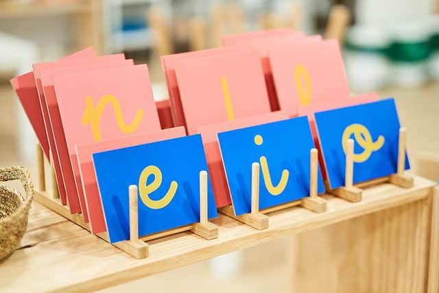 Photograph of sandpaper letters. The focus of the frame is three rectangular bright blue cards on which there is (L-R) a lowercase E a lowercase RINO lowercase O. These three cards are at an angle in the center of the frame. They are on a wooden table. Behind them are three larger cards on which there are(L-R) a lowercase and a lowercase L and an obscured letter. The litters appear to be yellow but they are made out of sandpaper.