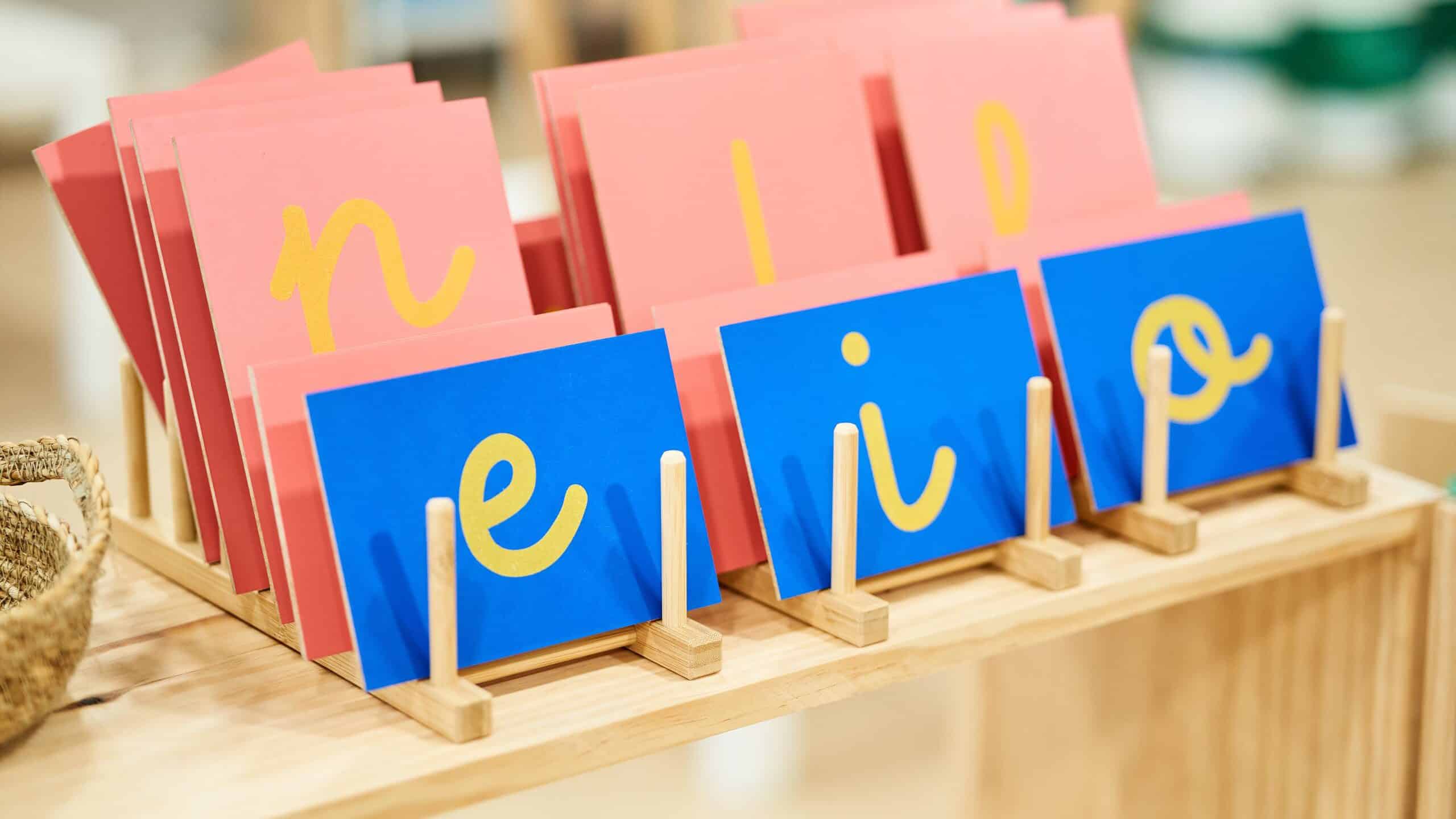 Photograph of sandpaper letters. The focus of the frame is three rectangular bright blue cards on which there is (L-R) a lowercase E a lowercase RINO lowercase O. These three cards are at an angle in the center of the frame. They are on a wooden table. Behind them are three larger cards on which there are(L-R) a lowercase and a lowercase L and an obscured letter. The litters appear to be yellow but they are made out of sandpaper.