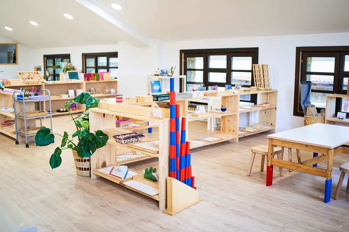 A photograph of a Montessori classroom. From the back left frame to the front there are two natural wood bookcases visible there is also a light blue metal rolling cart with three shelves visible in the back left frame. markers on the top shelf and then wooden trays on the second in lower shelf in front of the rolling cart is a plant in a wicker basket which is situated next to shelving that is at more of a vertical angle in the photograph. In the front right part of the frame there is a wooden table with red and blue feet and wooden stools. In the back of the frame there are four windows with dark frames visible. The windows each consist of nine separate panes of glass. The walls and the ceiling of the classroom are white. The floor is light natural wood. It is a very bright classroom