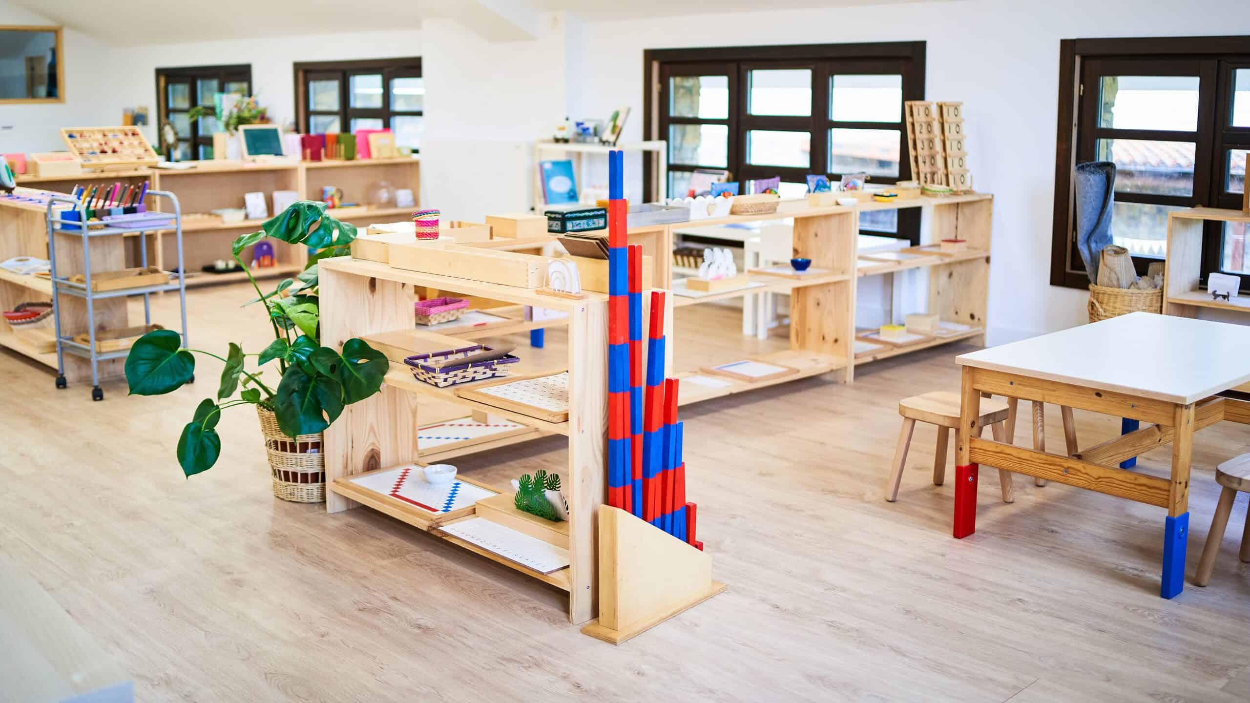 A photograph of a Montessori classroom. From the back left frame to the front there are two natural wood bookcases visible there is also a light blue metal rolling cart with three shelves visible in the back left frame. markers on the top shelf and then wooden trays on the second in lower shelf in front of the rolling cart is a plant in a wicker basket which is situated next to shelving that is at more of a vertical angle in the photograph. In the front right part of the frame there is a wooden table with red and blue feet and wooden stools. In the back of the frame there are four windows with dark frames visible. The windows each consist of nine separate panes of glass. The walls and the ceiling of the classroom are white. The floor is light natural wood. It is a very bright classroom