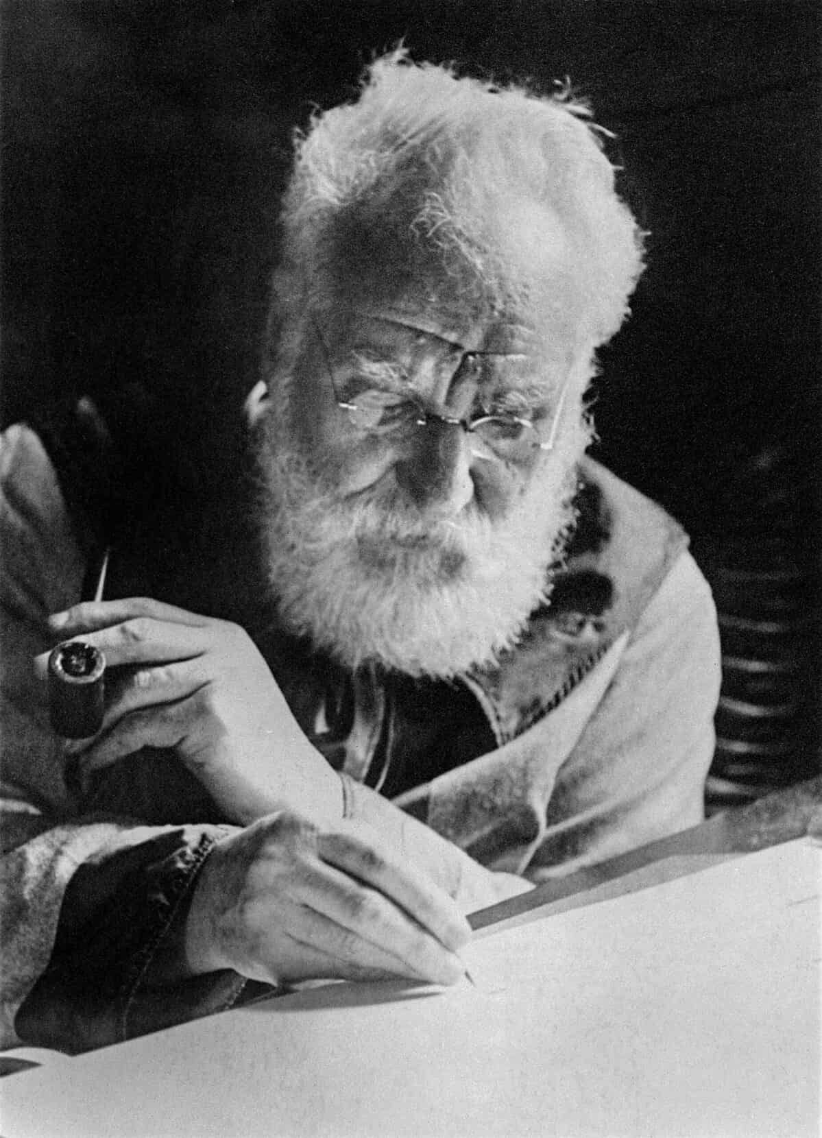 A black-and-white photograph of Alexander Graham Bell circa 1913. He is seated at his desk, which takes up the bottom and lower right frame. His gaze is focused looking down on the paper on which he writes with a pen in his right hand. In his left hand he is holding a pipe. His hair and his beard are gray and he is wearing wire-frame spectacles. He is quite wrinkled. indistinct black background. 