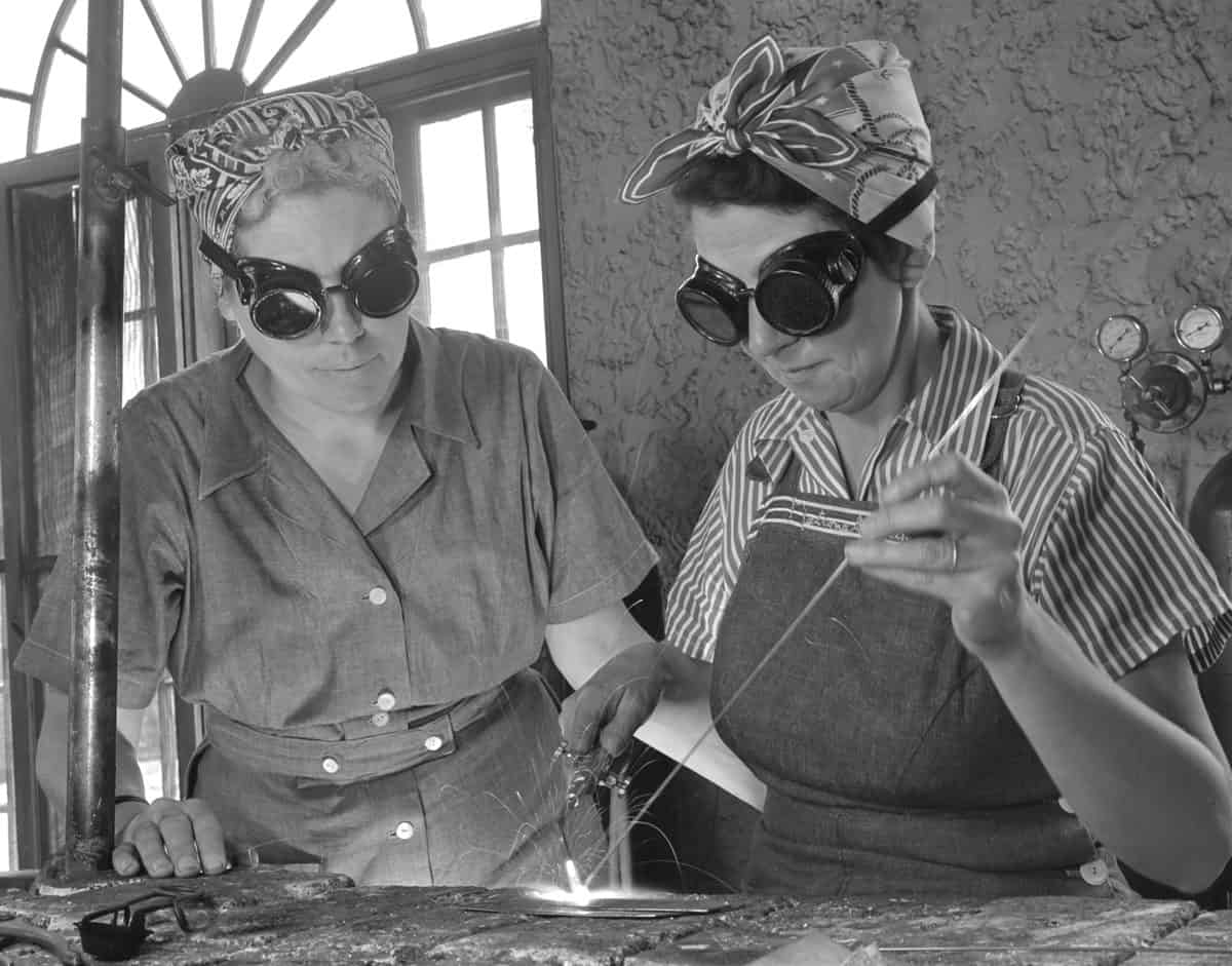 A black-and-white photograph from 1942 of two light-skinned women working in some sort of factory. The woman on the right side of the photograph is actively involved in soldering. She is wearing a short sleeve striped button front shirt with an apron over it, goggles over her eyes, and a scarf covering her hair. The woman on the left side of the frame is intently watching the woman on the right she is wearing a solid jumpsuit that buttons up the front. She is also wearing goggles over her eyes and a scarf covers her hair. The background is composed of some sort of tank with meters on it, right frame, and a window and possibly a door that is obscured by the woman on the left in the left background