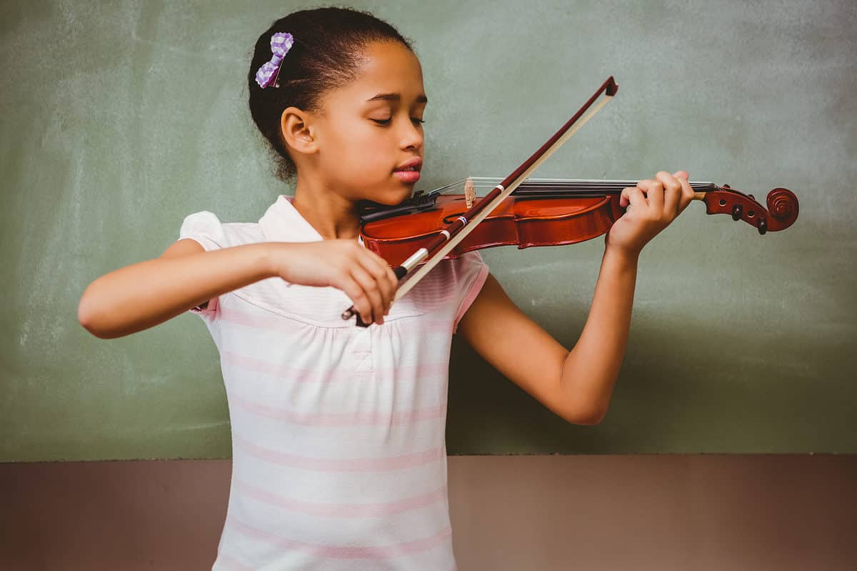 A young, slender, brown-skinned girl wearing a white and light pink striped top is visible slightly left of center frame playing a violin. Her dark hair is pulled back with a purple and white chick bow on the right side of her head her left arm is supporting the violin her right arm is holding a bow. Her gaze is focused on the violin. The background appears to be a chalkboard with nothing written on it.