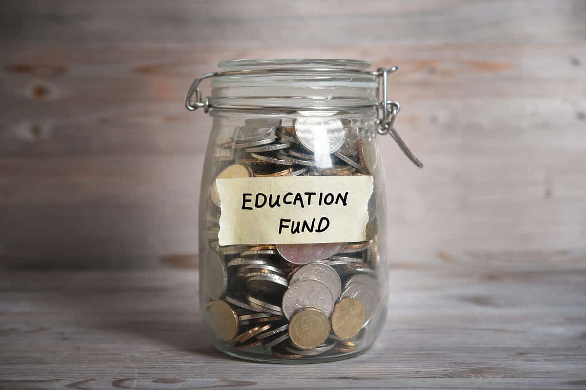 Center frame: A clear glass jar with a clear glass hinged lid is filled with mostly silver and a few gold coins. On a piece of masking that has been tor off the roll, are the words EDUCATION and FUND,. Education is above Fund. Neutral background.