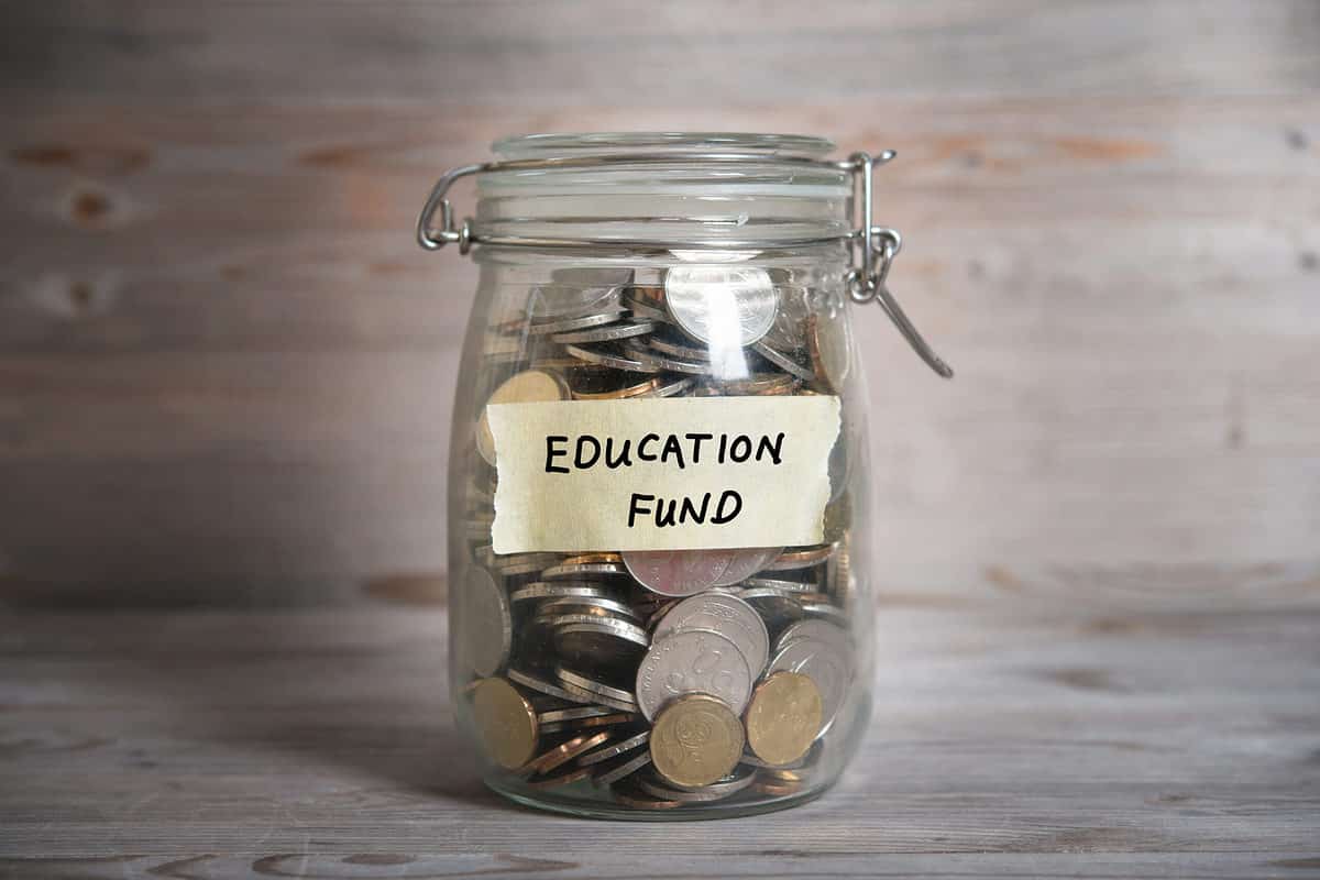 Center frame: A clear glass jar with a  clear glass hinged lid is filled with  mostly silver and a few gold coins. On a piece of masking that has been tor off the roll,  are the words EDUCATION and FUND,. Education is above Fund. Neutral background. 