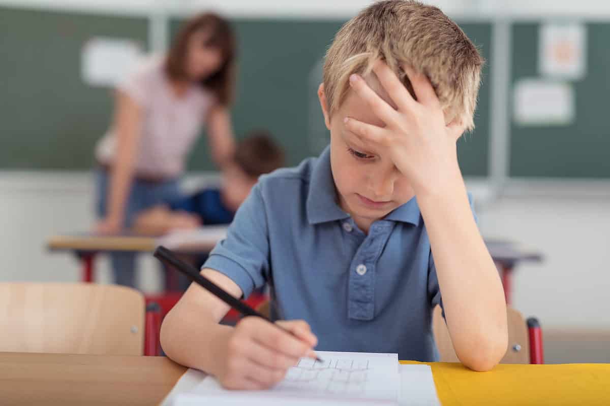 Center frame: a young malepreseting child is visible seated  at a desk. He is wearing a short-sleeved light blue polo-style shirt. He is holding a pencil in his right hand, while his left hand icon his forehead, as if propping it up. There are a couple odf sheets of white paper on the wooden desk.