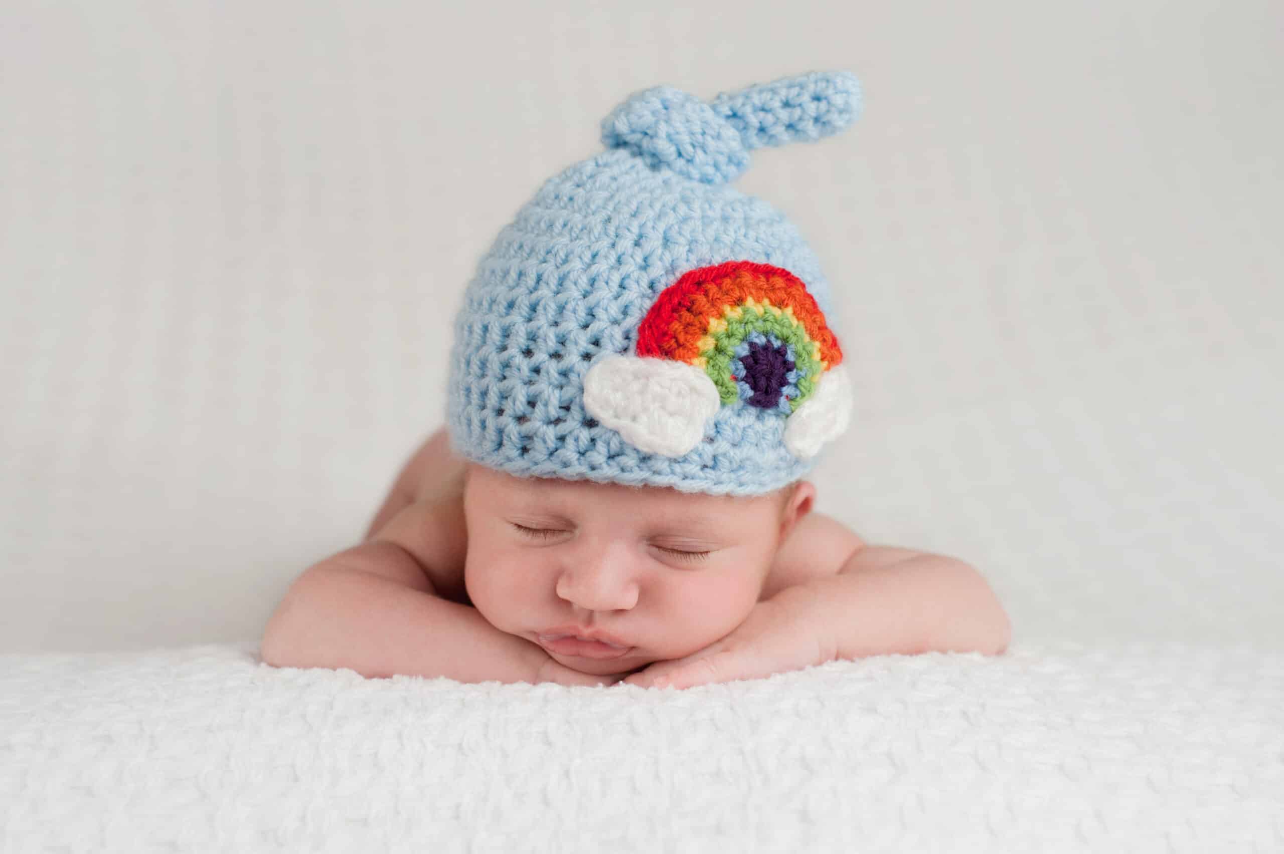 A sleeping, three-week-old, newborn baby boy wearing a top knot beanie with a rainbow design. Shot in the studio on a white blanket.