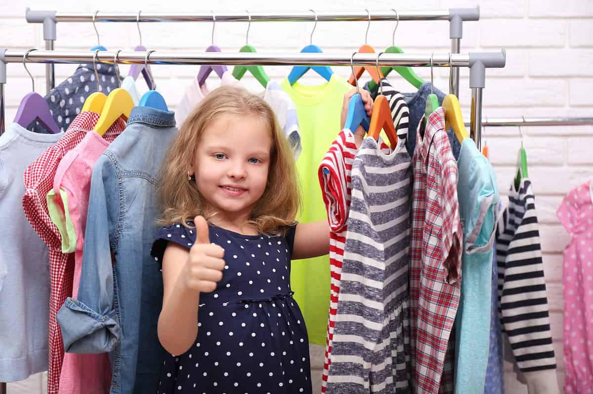 A light-skinned little girl, facing the camera and wearing a short-sleeved navy dress with polkadots Is visual center frame. She has shoulder length honey brown hair. She is standing directly Underneath a portable clothes hanging bar. She is standing between a number of different dresses on her left and right. Her left hand is holding a blue hanger from which hangs a red and white striped dress. Her right hand is out in front of her giving a thumbs up sign. From left frame the clothes are a gray shirt hanging from a purple wooden hanger, a red and white gingham dress hanging from a yellow wooden hanger a pink dress with a yellow him sleeve hanging from a yellow hanger, and a faded long sleeved denim dress hanging from a light blue hanger. To the little girls left are the red and white dress hanging from a blue hanger a gray and white striped dress hanging from an orange hanger a plaid short sleeved dress hanging from a Green Hanger. The background consists of more rods with clothes hanging from them however they are obscured by the little girl in the clothes in the front.