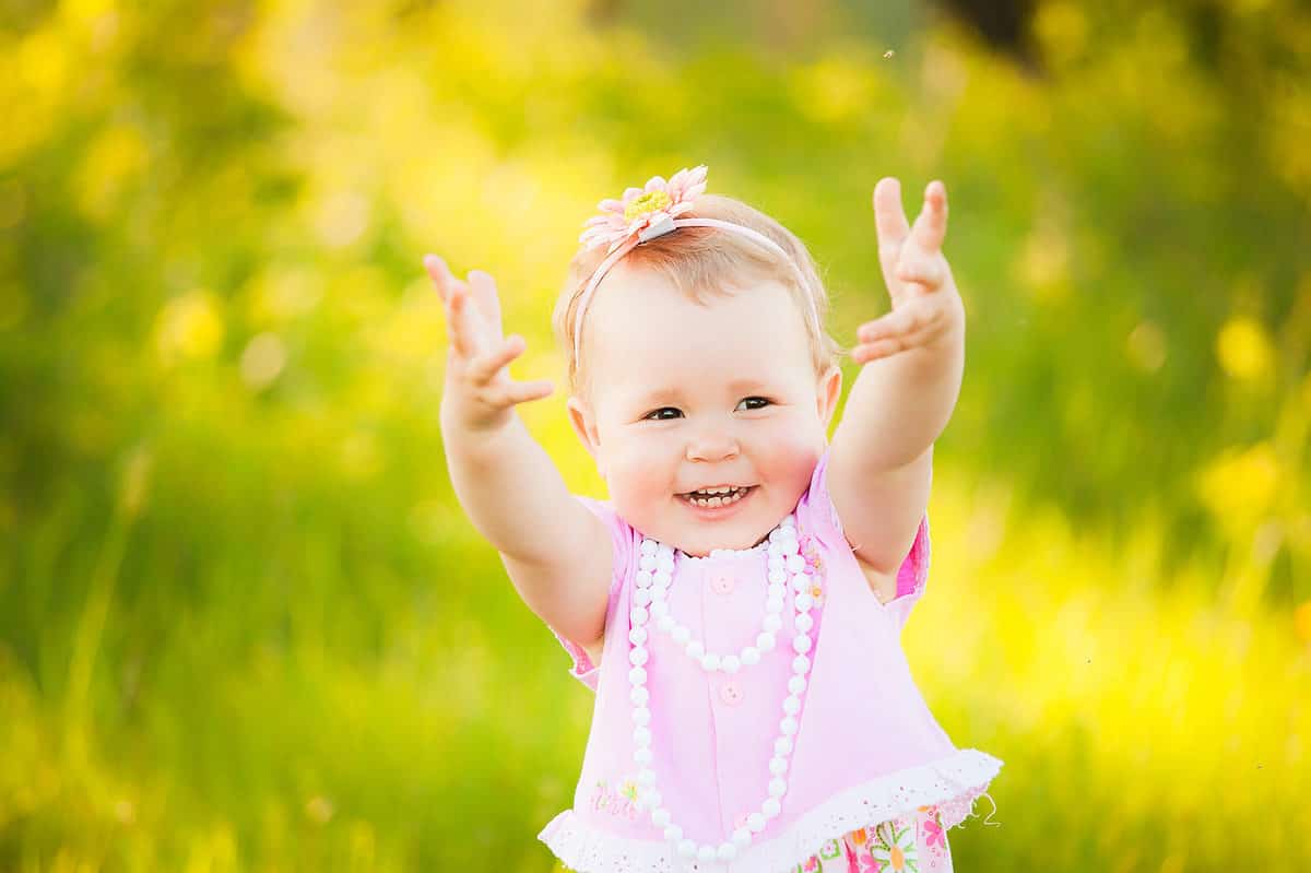 Portrait of happy joyful child in pink dress over wild yellow field flowers background. Family playing together outside. Mom and little daughter cheerfully blowing and catching soap bubbles.