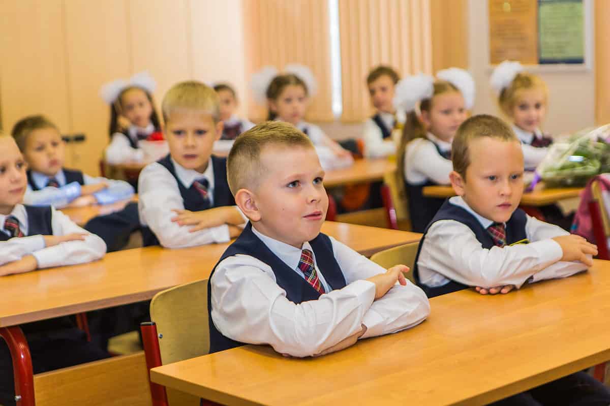 A male presenting child is visible center frame wearing a school uniform of a white long-sleeved button fromt collared shirt , over which is won a navy (blue0 sleeveless sweater vest. A tartan plaid tie is visible at the color. He is light-skinned . His arms are crossed in from of him on the desk that he shares with a mother student, right frame who looks remarkably similar to the first boy. Ten other similarly dressed light-skinned students are seated around them, but are not subject t the camera's focus. All of the children have their arms crossed in front of them. There are four female appearing students with white hair bows. 