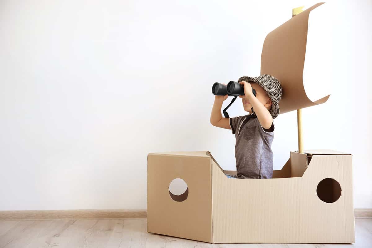 frame right: A young child wearing a gray short-sleeved t-shirt and a matching derby-style hat is holding binoculars up to his face as if looking at something in the distance. He is in a "boat" fashioned from a khaki-colored cardboard box which as a khaki-colored "sail" made from kraft paper impaled on a dowel. The back ground is a white wall. The "boat" is on a wooden floor.