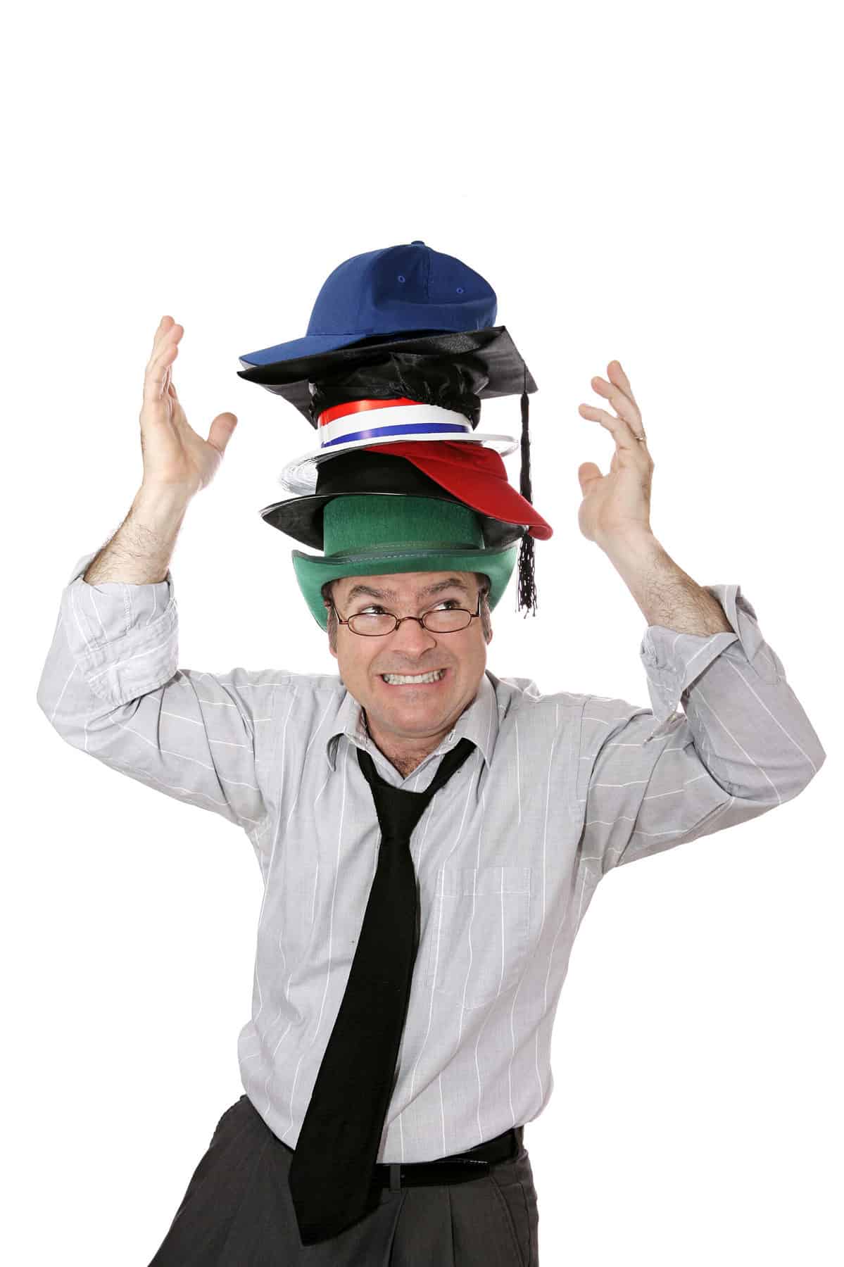 A light-skinned, male-presenting adult wearing a gray with slender white vertical stripes, long-sleeved button from collared shirt, unbuttoned at the collar, with a loosened dark tie . He is wearing eye glasses. He has seven hats of various colors on his head. He is holding his arms /hands up in a gesture that implies he is concerned that the hats might fall. White isolate background.