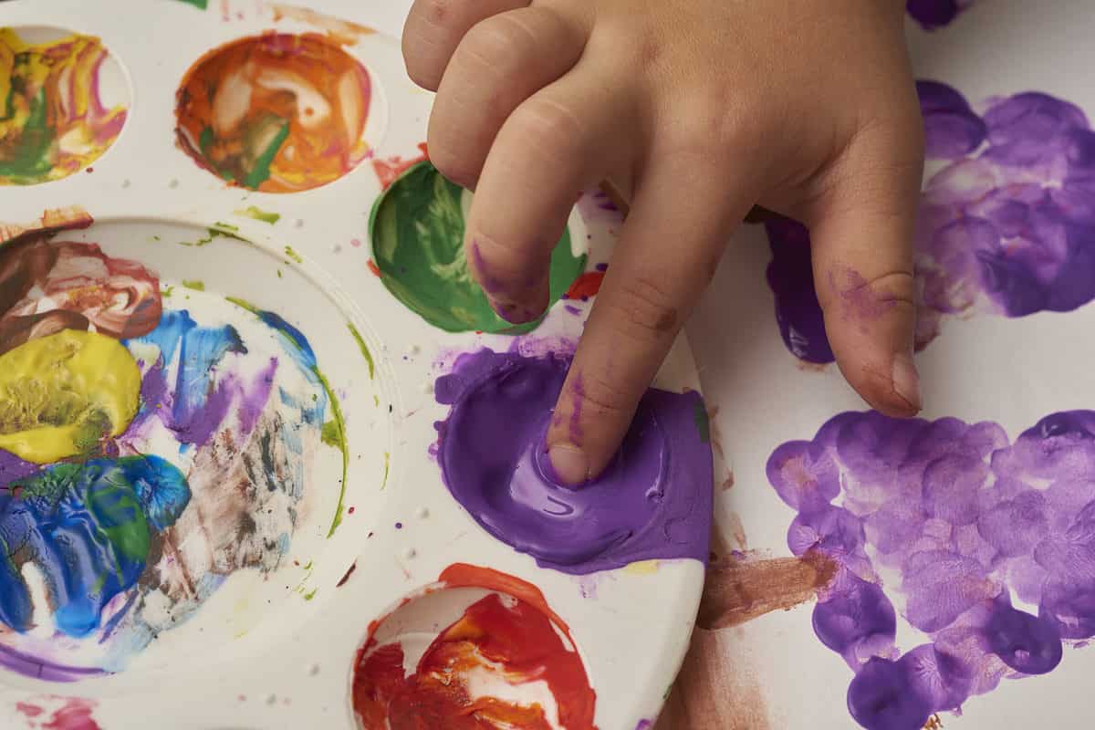 A child's right hand, upper right frame, is visible dipping index finger in purple paint. Thepaint is in a white round palette at 3 o'clock.Practically empty orange  paint is on the palette at 1 o'clock, with green at 2 o'clock, and red at 4 o'clock.