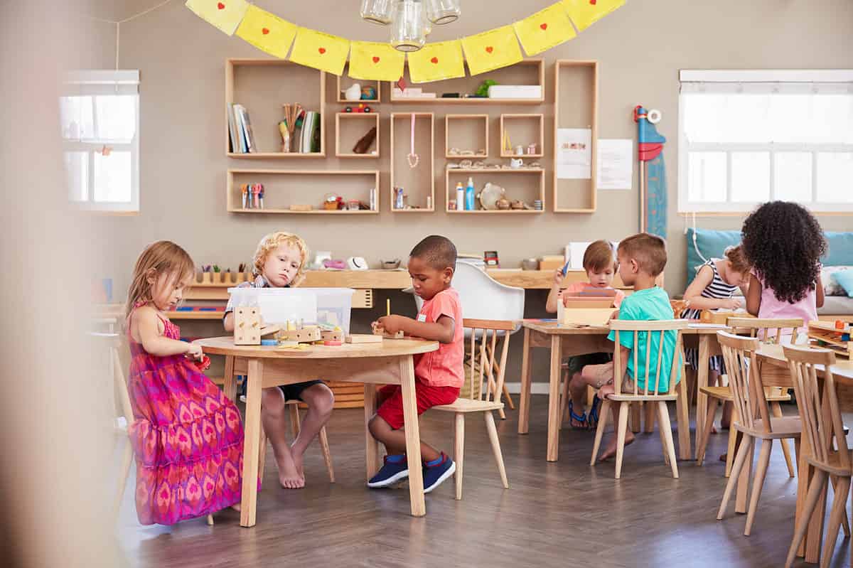 A classroom with a wooden laminate floor. The background is a neutral colored wall on which hang shelves with books and glue and markers, in the renter frame. Windows are in the wall on the left and right frames.  In the classroom itself, four tables. There are three children seated At the table and frame left closest to the left is a light-skinned little girl with stringy light brown hair. She is  wearing a pink and orange sundress. She is looking at some things she is holding in her hand bit away from the table. To her left closer to the center of the frame is a light-skinned little boy with short blonde hair;  he is obscured by a plastic tub that is on the table in front of him, but underneath the table you can see that he is wearing shorts and he is barefoot. Seated to his left in the center of the frame is a brown skin boy with very close cropped black hair he is wearing a short-sleeved coral colored T-shirt and rust colored shorts he also has on black tennis shoes with white soles. He has a pencil in his left hand or possibly a wooden dowel. Behind them are four other children two little boys and two little girls. A light-skinned boy facing the camera is looking down at something on the table. He is obscured by a basket on the table. Across the table from him with his back to the camera is a olive-skinned little boy wearing a short sleeve awkward T-shirt and khaki shorts he is sitting crosslegged in the chair. A light-skinned little girl in the back right frame wearing a blue and white striped sundress has her head down performing a task at the table. A brown-skinned girl with waist long black hair has her back to the camera across the table from the little girl wearing the sundress