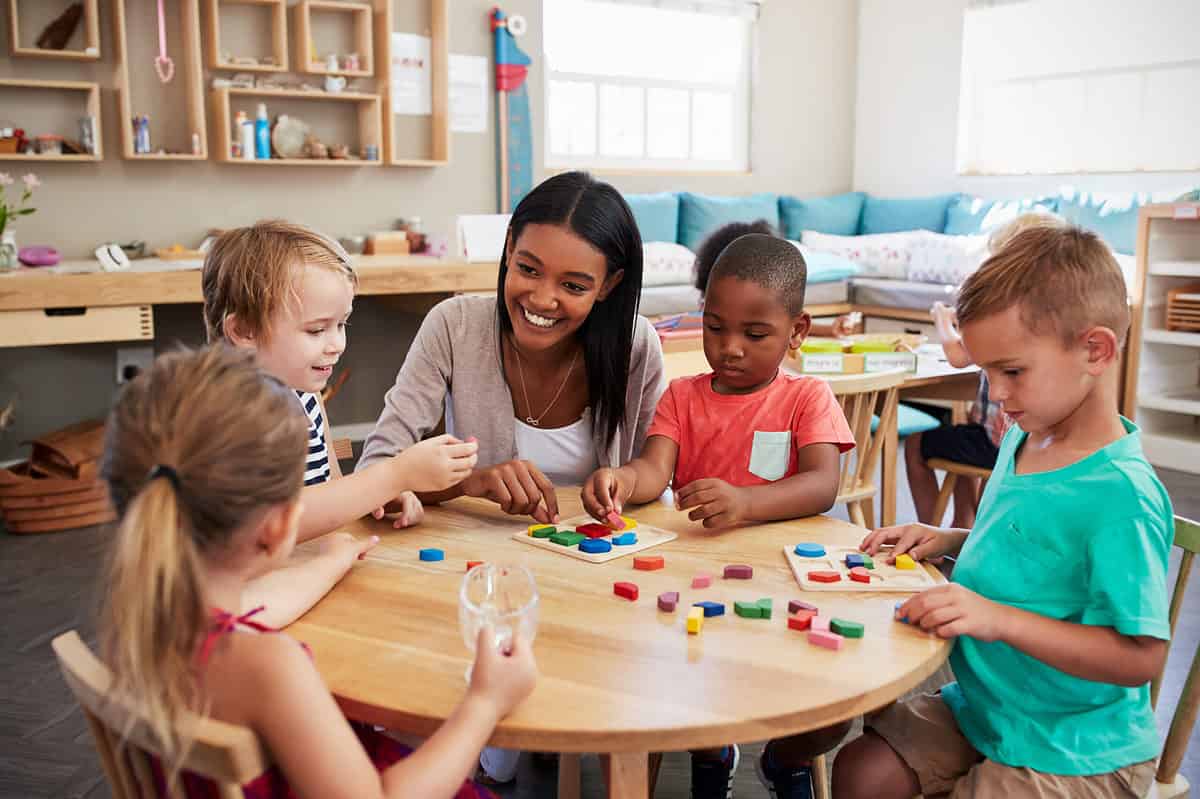 A brown-skinned smiling female presenting adult with long dark hair, wearing a white scooped-neck t-shirt and a beige sweater is visible crouching at a low round, wooden table with 4 children: a brown-skinned, male-presenting child wearing a coral-colored short-sleeved t-shirt with a white pocket on the left breast. He id =s to the teacher's left, but to her right in the frame. Next to that kid, is an olive-skinned male presenting child wearing an aqua sport sleeved t-shirt. Next to him, front left frame a female =presenting child is visible with her back toward the camera. She is light-skinned, her hair is light-brown and pulled up in an unremarkable elastic. A light=skinned, male presenting child sits next to her, upper left from. He is wearing a navy and white striped shirt. All of the people in the photo are focused on bright colored manipulatives small wood shapes used in math activities.