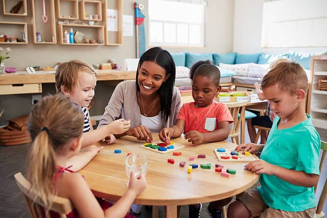 A brown-skinned smiling female presenting adult with long dark hair, wearing a white scooped-neck t-shirt and a beige sweater is visible crouching at a low round, wooden table with 4 children: a brown-skinned, male-presenting child wearing a coral-colored short-sleeved t-shirt with a white pocket on the left breast. He id =s to the teacher's left, but to her right in the frame. Next to that kid, is an olive-skinned male presenting child wearing an aqua sport sleeved t-shirt. Next to him, front left frame a female =presenting child is visible with her back toward the camera. She is light-skinned, her hair is light-brown and pulled up in an unremarkable elastic. A light=skinned, male presenting child sits next to her, upper left from. He is wearing a navy and white striped shirt. All of the people in the photo are focused on bright colored manipulatives small wood shapes used in math activities.