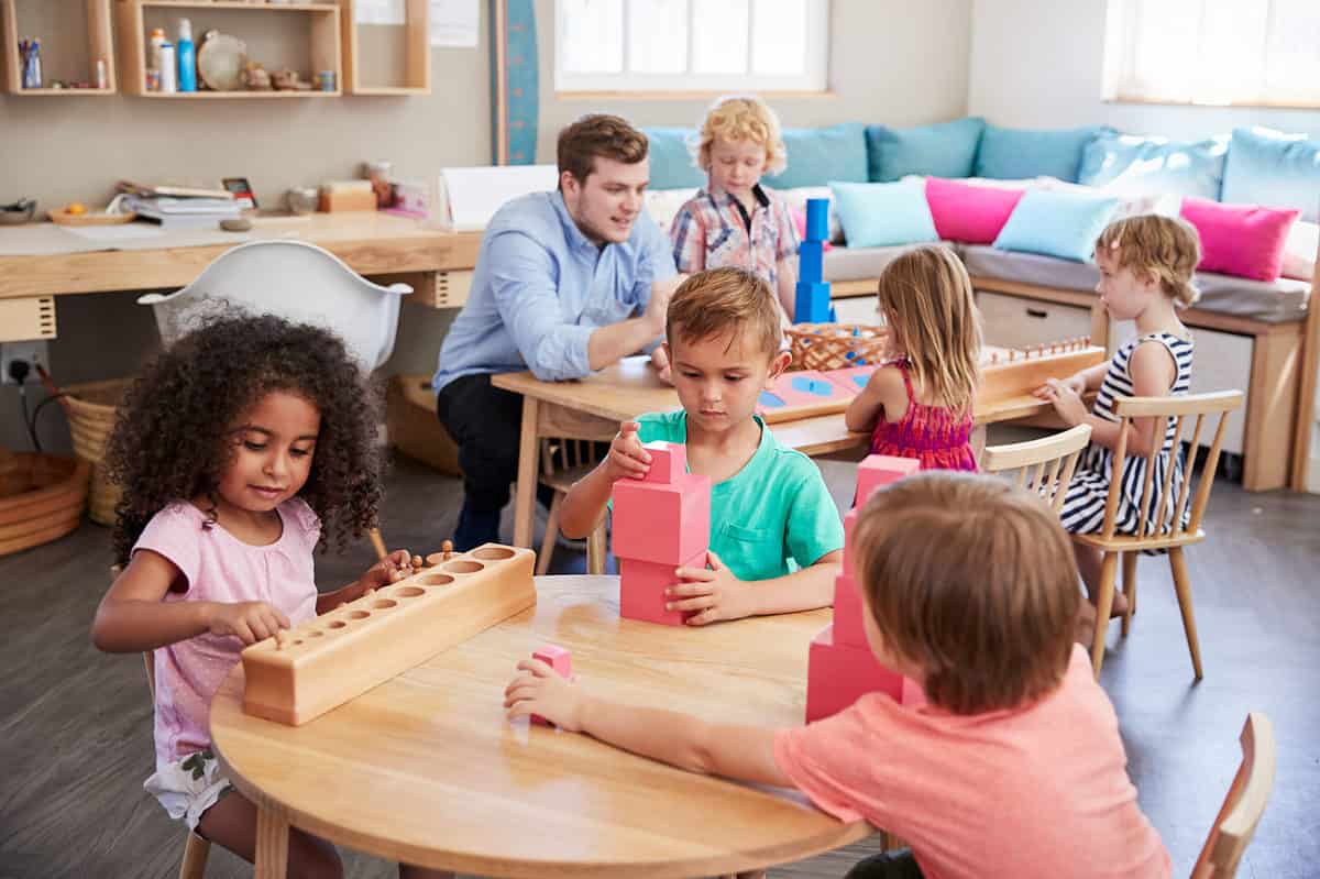 Full frame of a Montessori classroom toward the front of the frame is a round wooden table at which three young children are seated. A young brown-skinned girl with black curly hair wearing a shirt-sleeved pink t-shirt toward the left part of the frame seated at the table which is child-sized, playing with a wooden block with graduated holes the smallest being toward the front of the frame getting increasingly larger as you go towards centerpoint. A light-skinned male child With light brown hair wearing a seafoam green short-sleeved T-shirt is facing the camera and playing with three pink blocks that he has stacked. Frame right is the back of a light-skinned boy whose left arm is extended across the table reaching for a pink block he is wearing a shirt-sleeved salmon colored T-shirt. In the back of the frame arethree children and an adult man seated at a rectangular child sized table. The adult male is hunched down leaning against the table using his elbows for support, next to him is a light skin boy with longish blonde hair wearing a button front shirt that is plaid mostly red white and blue. He is looking down at some thing the teacher is doing but is obscured by the head of the child wearing the green shirt. Across the table from the teacher and the blonde haired boy are two little girls whose backs are facing the camera the girl on the left is wearing a sundress that is primarily magenta with orange accents the girl on the right is wearing a navy and white striped dress. The background consists Of an L-shaped sofa with blue and pink pillows, Upper right frame and then the upper left frame an adult size desk with a Charles Eames chair. The chair is white.