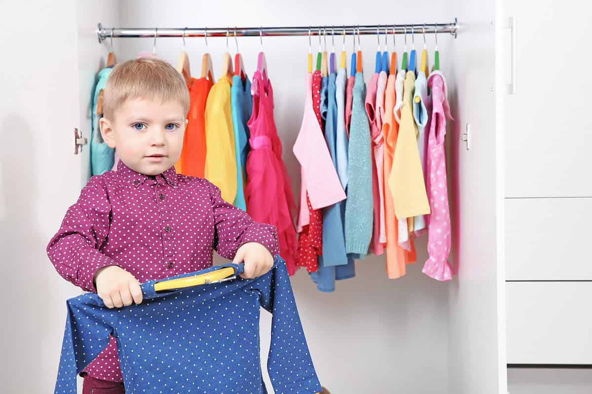 A light skin a little male presenting child with short blonde hair is visible in the left frame of the photo. He is wearing a long-sleeved burgundy polkadot button front shirt. In his hands in front of him he is holding a blue polkadot shirt on a wooden hanger. Behind him is an white open closet with a silver metal rod upon which many other clothes hang. Most of the clothes in the closet appear to be what are traditionally thought of as female presenting. From left to right there is a seafoam green down vest. The next two items of clothing are obscured by the little boy’s head. To the right of his head or long sleeve orange yellow and blue shirts. There is a magenta fancy cocktail dress next to the blue shirt. Then there is a space in the clothing. From left to right the next group of clothing is a long sleeve yellow shirt a red polkadot button front shirt a short sleeve blue shirt a long sleeve blue shirt very light blue long sleeve shirt a coral color some thing that’s obscured by a long sleeve blue heather sweater. To the right of the blue heather sweater or pink light pink coral white short sleeve shirts and a yellow long sleeve shirt a light blue short sleeve shirt and a pink polkadot romper