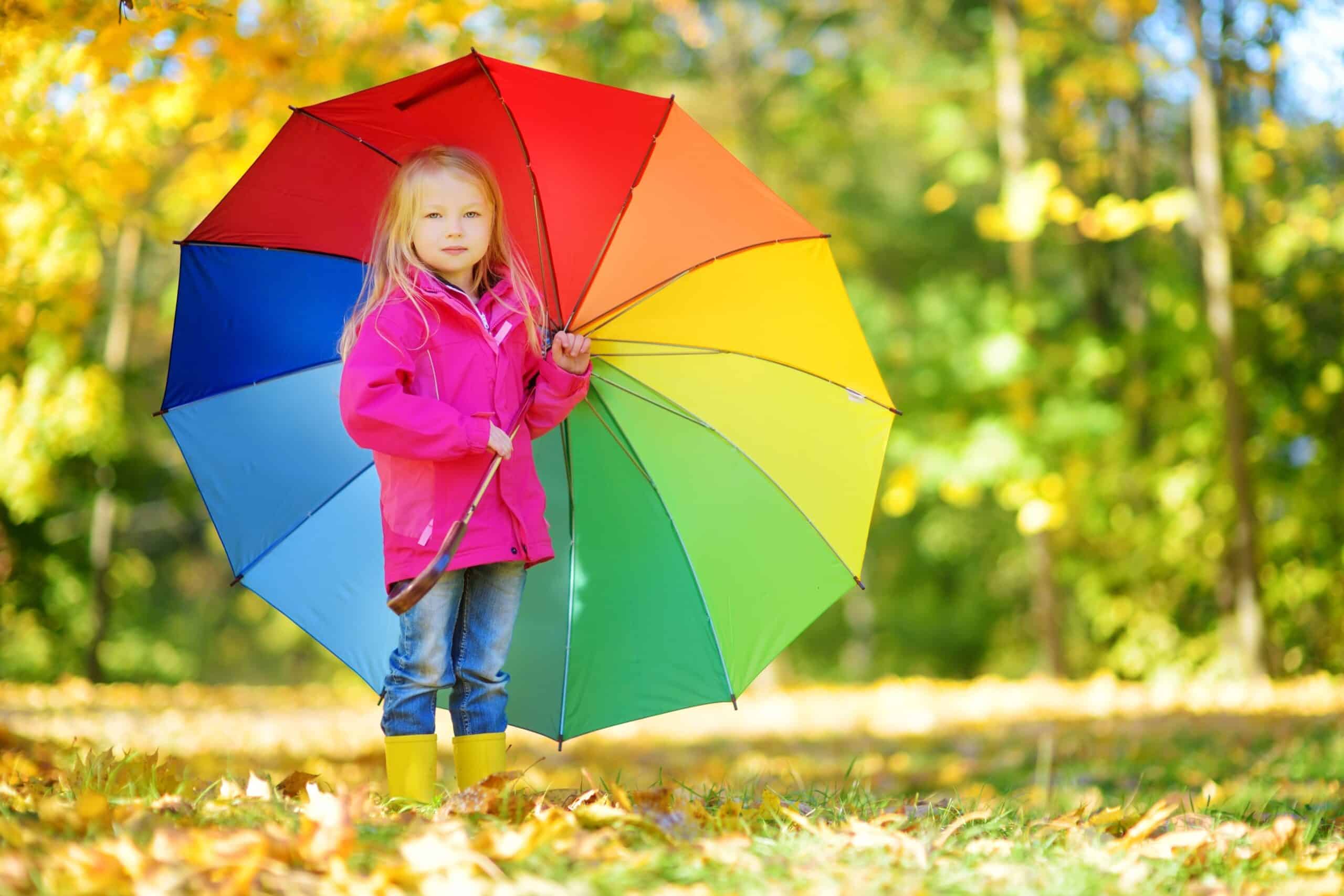 Cute little girl holding rainbow umbrella on beautiful autumn day. Happy child playing in autumn park. Kid gathering yellow fall foliage. Autumn activities for children.