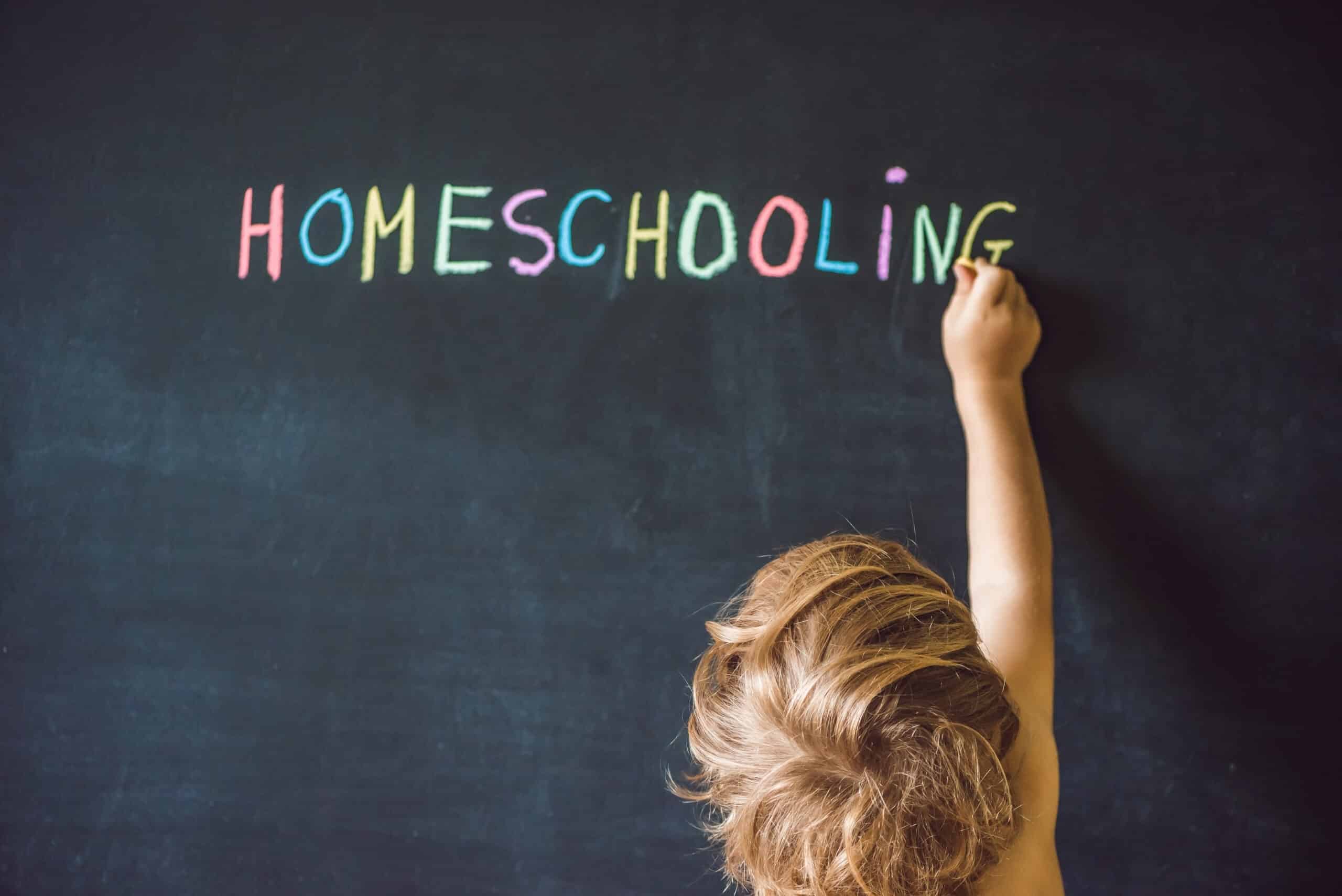 A light-skinned child with honey-brown hair, bottom center/right frame, facing away from the camera is visible holding a piece of chalk in their right hand, up tho a chalkboard as if finishing writing the word HOMESCHOOLING. HOMESCHOOLING is written on a blackboard which also serves as the background. The letters in HOMESCHOOLING are written in different colors - H,S, the third O, and I are pink, The first O, C, and L are blue. The E, the second O, and the N are light green/white, and the M,H, G are yellow.