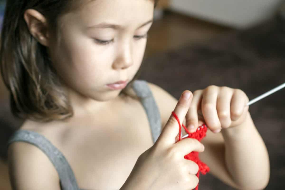Full frae:Young female-presenting child with asian features is concentrating on knitting with red yarn. She has a strand of yar looped around the index finger of her right hand.