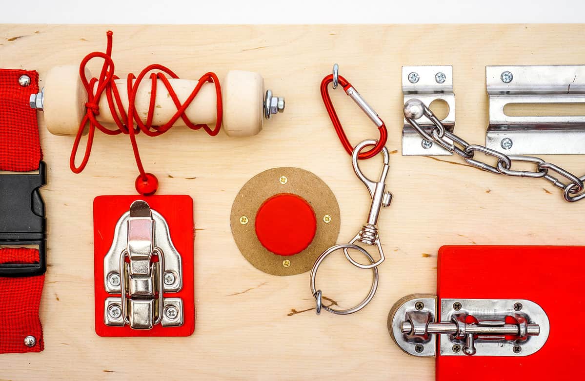 Full frame photograph a “busy board,” consisting of a clasp, a latch a surface bolt, and a chain lock. The board itself is natural color pieces of wood attached to the board are red and most of the hardware is silver.