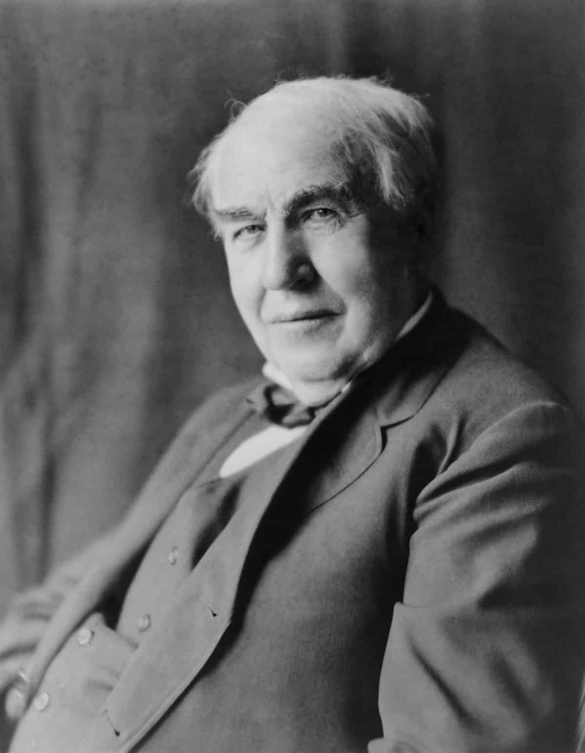 A black-and-white portrait of Thomas Edison. He is facing the left side of the frame that he is looking forward. He is an older man with male pattern baldness and scraggly gray hair though his bushy eyebrows appear somewhat brown. He is wearing a three-piece charcoal suit the jacket is open but the vest is buttoned. He is a bit on the rotund side. He’s got a Mona Lisa smile.