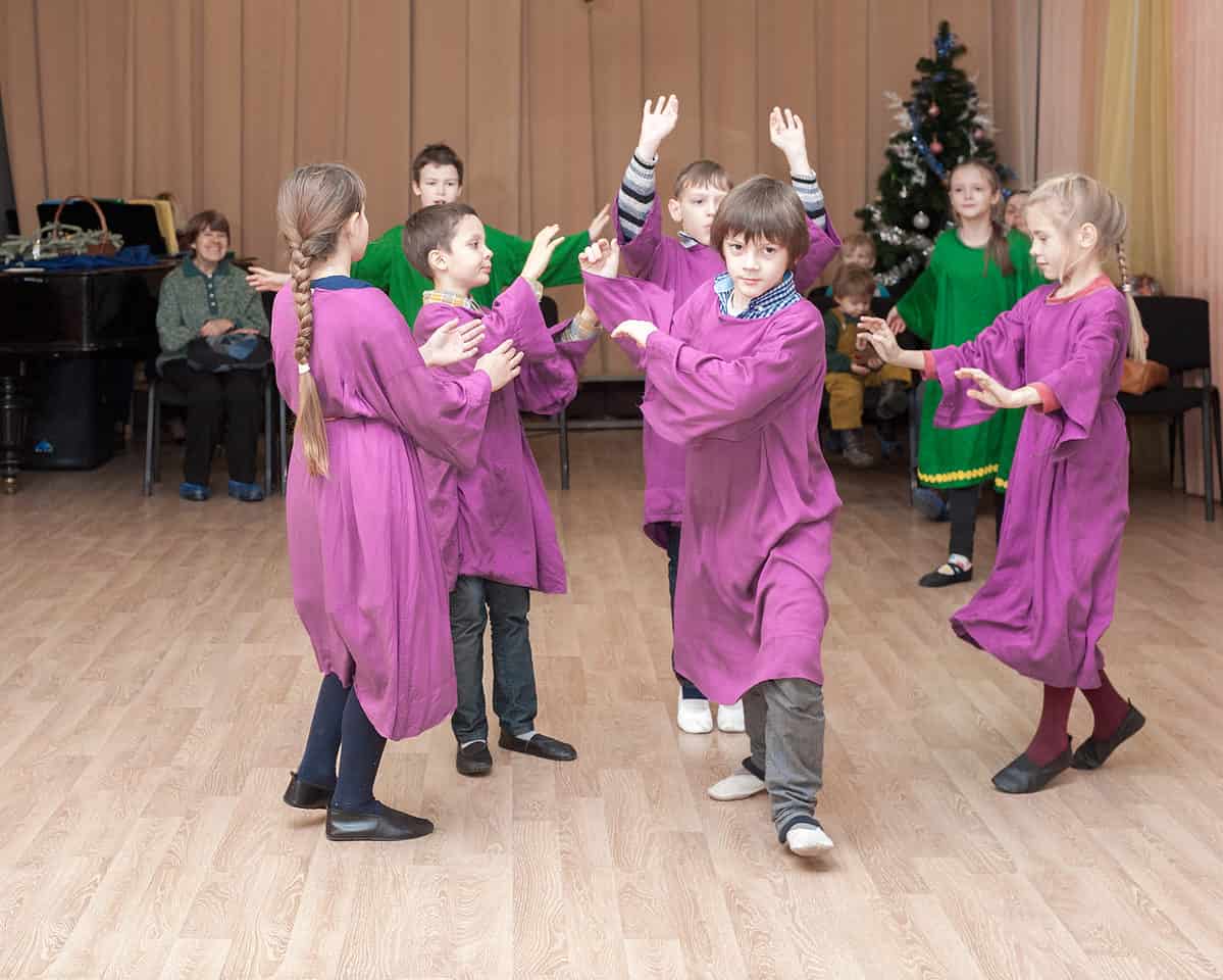 Center frame:Two children dressed in green eurythmy robes are visible behind five Waldorf students, moving in a clock-wise circle, dressed in purple eurythmy robes. An adult, left background is visible smiling as if pleased by the children's performance. A Christmas tree is evident in the right background. 