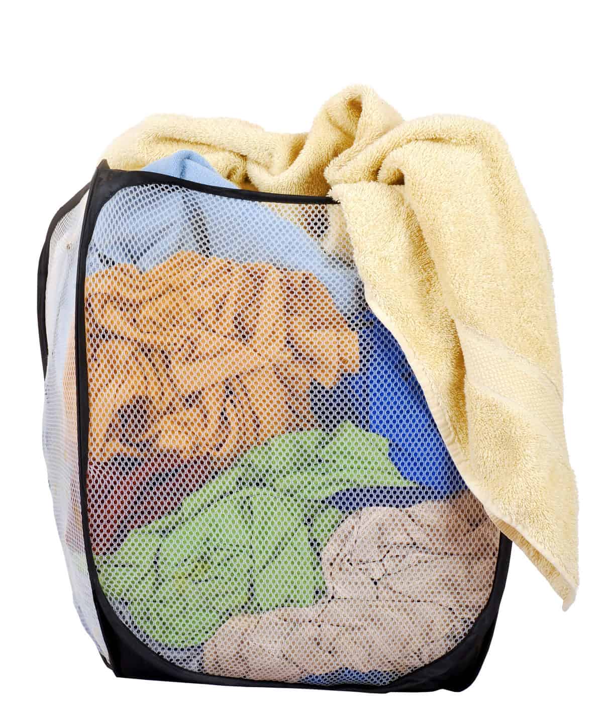 Photograph of a white mesh clothes hamper with black trim. The hamper is full of cloth/clothing of a discernible type from the bottom to the top there is some light beige cloth on top of which is some sage green royal blue there’s some maroon cough and then a lot of gold cloth and then hanging out of the hamper is a bath towel that is wheat colored or light yellow. Photograph is sit against white isolate.