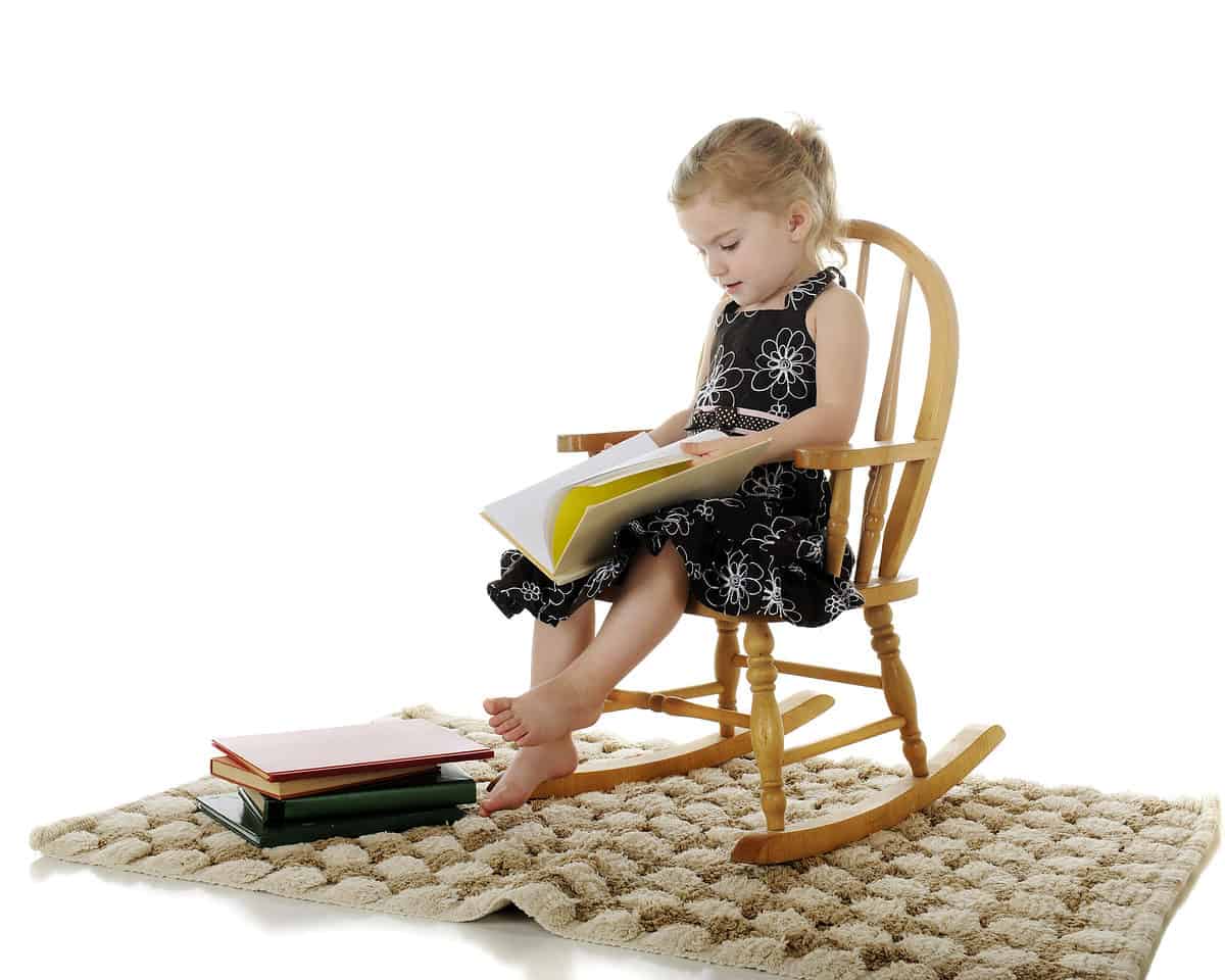 A photograph of a light-skinned little girl in a sleeveless sundress that is either black or navy with white outlines of flowers on it and a polkadot belt is visible sitting in a child-sized wooden rocking chair on a jute of sisal rug. She is barefoot. In her lap is a white book that is open but appears to have blank pages. At her right foot on a rug, four books are haphazardly stacked. Against white isolate background