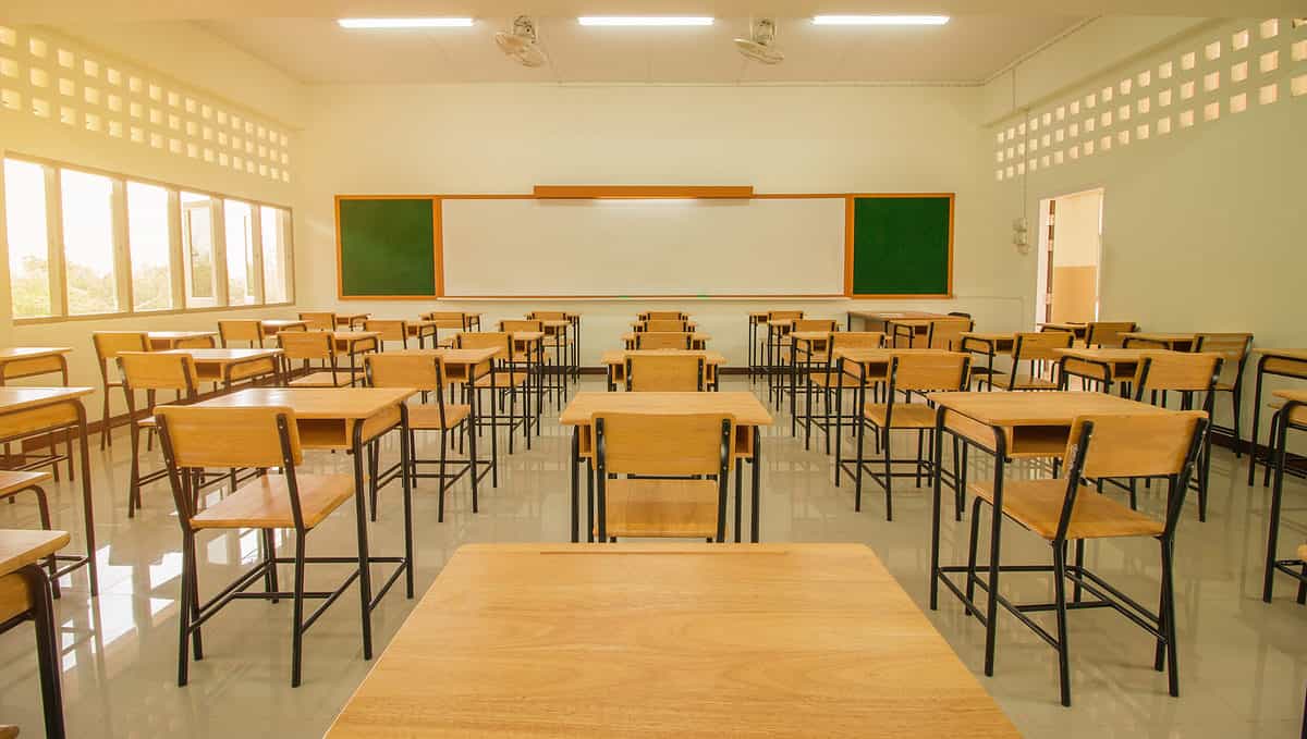 A classroom with 5 rows of natural wooden desks and chairs with slender black metal legs. The photograph is taken from the back of the room. The back of the frame is a rectangular white dry erase board with a square green chalk board on its left and right. Three skylights are visible at the top the back frame. 