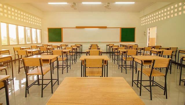 A classroom with 5 rows of natural wooden desks and chairs with slender black metal legs. The photograph is taken from the back of the room. The back of the frame is a rectangular white dry-erase board with a square green chalkboard on its left and right. Three skylights are visible at the top of the rear rack.
