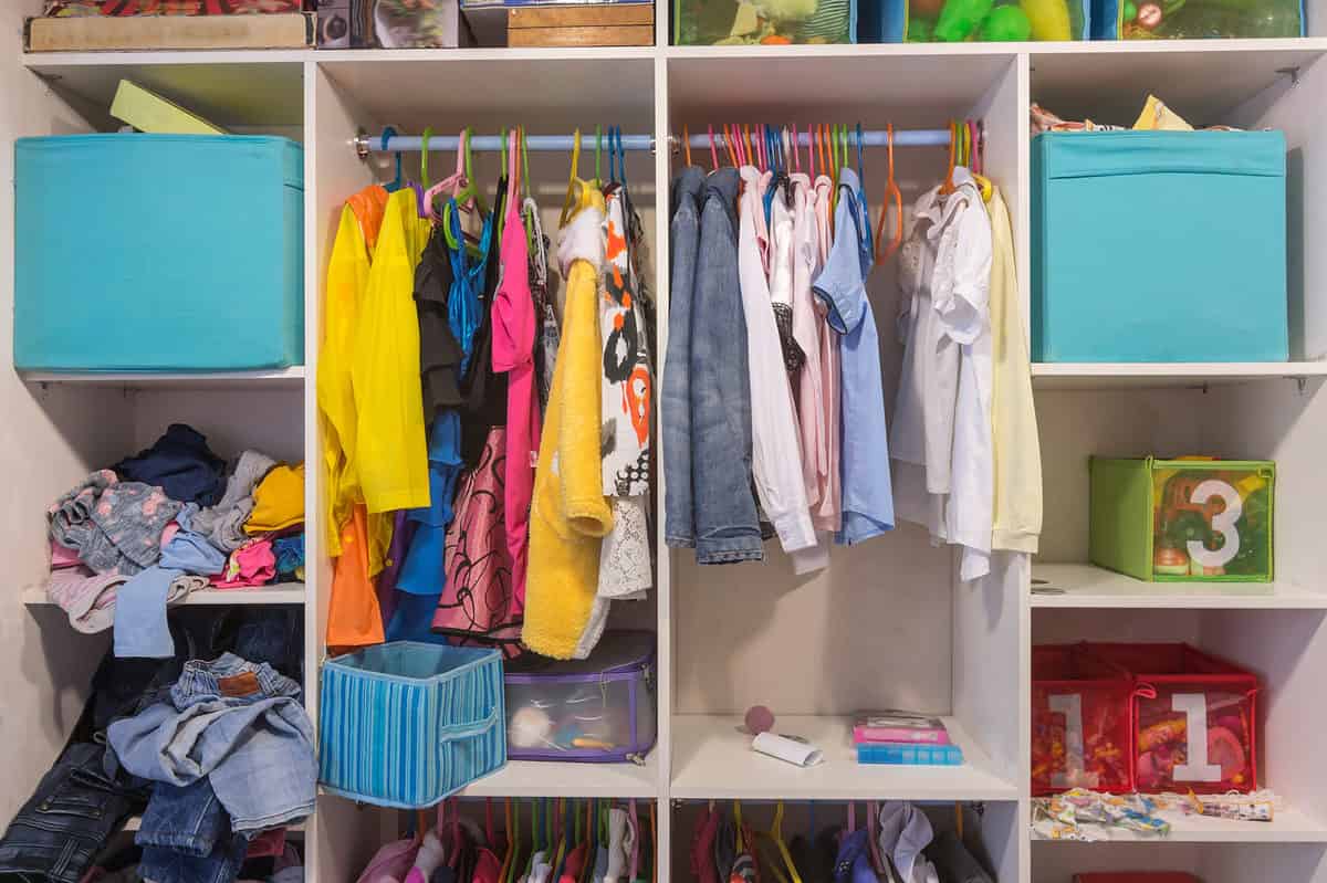 Full frame photo of a child’s open white wood wardrobe. On the left side of the wardrobe is a aqua blue fabric cube in the top shelf below it is a shelf it looks like clothes have just been shoved in it from a laundry basket. Those clothes appear to be mostly shirts. The shirts are blue yellow pink and one that is gray and pink. In the queue below the shirts are three pairs of blue jeans visible that look like they’ve kind of just been haphazardly put away as well there is something below the jeans but it is not visible in the photograph. To the right of those shelves is a large rectangular cube with a rod. There are 13 hangers on the rod. The hangers are blue pink green orange and yellow. The clothes in the left cube look like costumes in this particular Though there may be a kid-sized cocktail dress, but it’s more likely a princess costume. There is a terry cloth robe, too, but that may in fact be a “The Dude” costume. On the shelf beneath the rod is a blue fabric cube with a handle it appears to be empty though there may be things lower down in it behind the fabric cube is a mesh cube with a purple rim that has some thing indiscernible in it In the cube to the right of that cube there are 20 or so hangers upon which regular looking clothing is hanging, primarily shirts and a couple of jackets. The jackets are grayish powder blue. A long sleeve white shirt hangs to the right of the jackets. a short sleeve pink shirt hangs next to the white shirt. There are a indiscernible pieces of clothing between the short sleeve pink shirt and then a short sleeve blue shirt. Directly to the right of the blue shirt is an empty orange hanger. To its right is a short sleeve white blouse and then another short sleeve white blouse and then an empty yellow hanger and then a yellow blouse. On the shelf underneath the clothes are some indiscernible objects- possibly books and maybe a ball. In frame right are three shelves the top shelf has an aqua fabric cube with some things sticking out that are in describable below that is a mesh front cube with a three stenciled onto it. It is green and seems to have toys in it below that are two red mesh cubes that have the number one stenciled on each of them. Not sure what they contain. There is some cloth in front of them on the shelf. In the bottom half of the frame is actually another lower shelf and rods, but it is out of the frame for the most part.