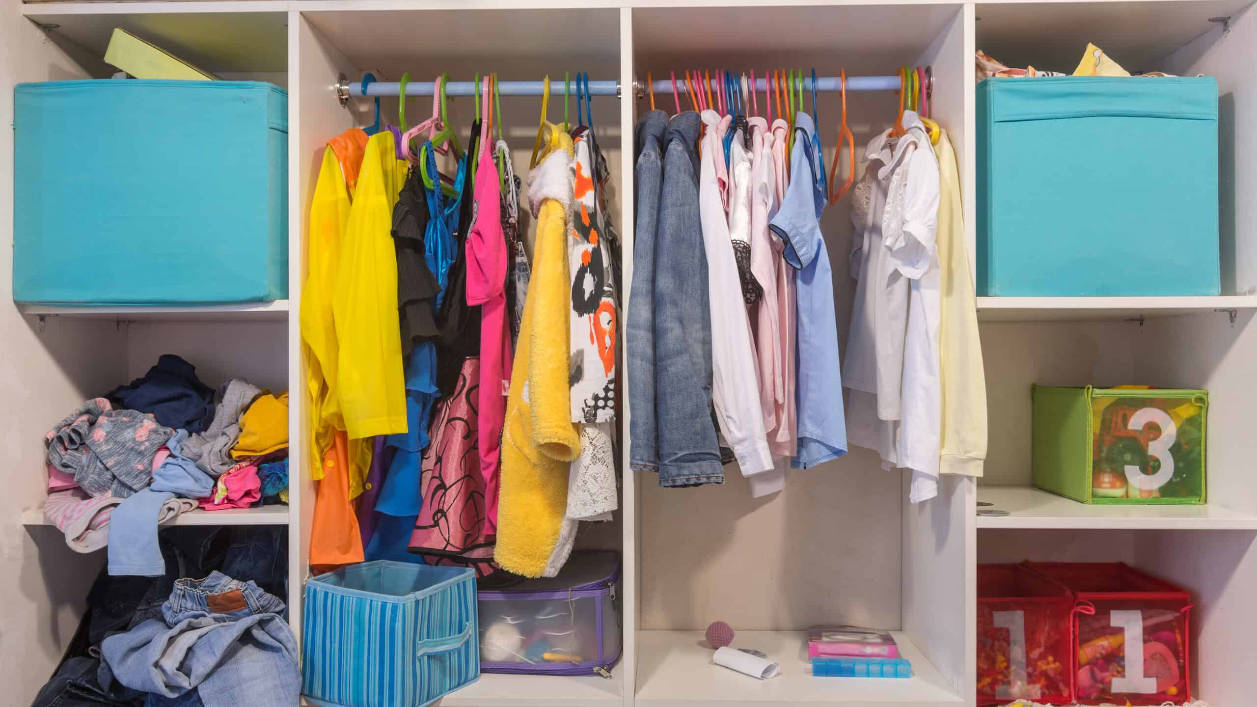 Full frame photo of a child’s open white wood wardrobe. On the left side of the wardrobe is a aqua blue fabric cube in the top shelf below it is a shelf it looks like clothes have just been shoved in it from a laundry basket. Those clothes appear to be mostly shirts. The shirts are blue yellow pink and one that is gray and pink. In the queue below the shirts are three pairs of blue jeans visible that look like they’ve kind of just been haphazardly put away as well there is something below the jeans but it is not visible in the photograph. To the right of those shelves is a large rectangular cube with a rod. There are 13 hangers on the rod. The hangers are blue pink green orange and yellow. The clothes in the left cube look like costumes in this particular Though there may be a kid-sized cocktail dress, but it’s more likely a princess costume. There is a terry cloth robe, too, but that may in fact be a “The Dude” costume. On the shelf beneath the rod is a blue fabric cube with a handle it appears to be empty though there may be things lower down in it behind the fabric cube is a mesh cube with a purple rim that has some thing indiscernible in it In the cube to the right of that cube there are 20 or so hangers upon which regular looking clothing is hanging, primarily shirts and a couple of jackets. The jackets are grayish powder blue. A long sleeve white shirt hangs to the right of the jackets. a short sleeve pink shirt hangs next to the white shirt. There are a indiscernible pieces of clothing between the short sleeve pink shirt and then a short sleeve blue shirt. Directly to the right of the blue shirt is an empty orange hanger. To its right is a short sleeve white blouse and then another short sleeve white blouse and then an empty yellow hanger and then a yellow blouse. On the shelf underneath the clothes are some indiscernible objects- possibly books and maybe a ball. In frame right are three shelves the top shelf has an aqua fabric cube with some things sticking out that are in describable below that is a mesh front cube with a three stenciled onto it. It is green and seems to have toys in it below that are two red mesh cubes that have the number one stenciled on each of them. Not sure what they contain. There is some cloth in front of them on the shelf. In the bottom half of the frame is actually another lower shelf and rods, but it is out of the frame for the most part.