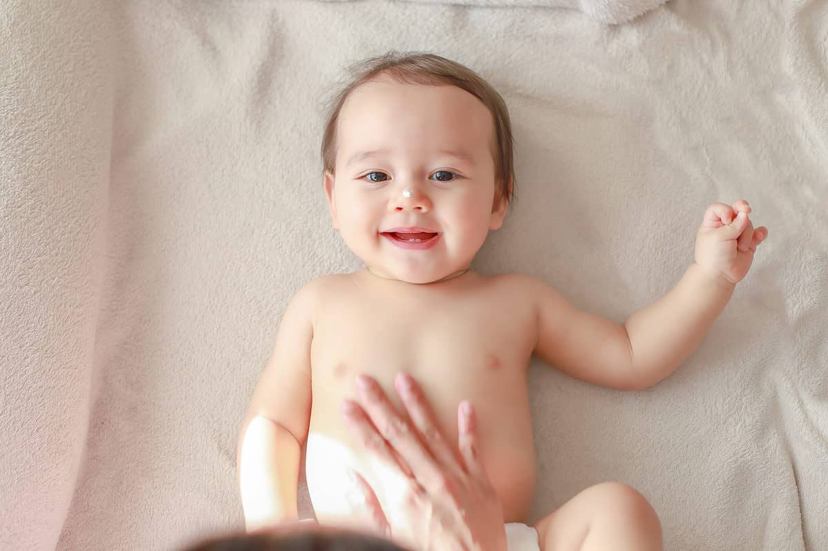 Cute baby boy applying lotion cream on face and body by his mommy making massage after bathing. Newborn child temporary first milk teeth smiling face top view. Mixed race Asian-German kid.