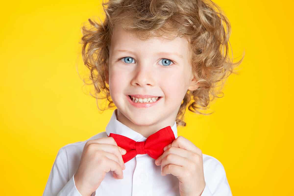 Portrait of a curly-haired European boy with blue eyes in a shirt and a red bow tie on a yellow background. A Slavic type. The child is laughing, smiling, having fun, happy.