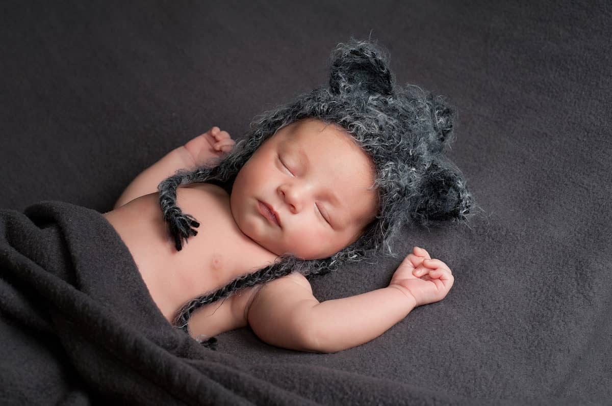 Three-week-old newborn baby boy wearing a gray crocheted wolf hat. He is sleeping on his back on a gray blanket.