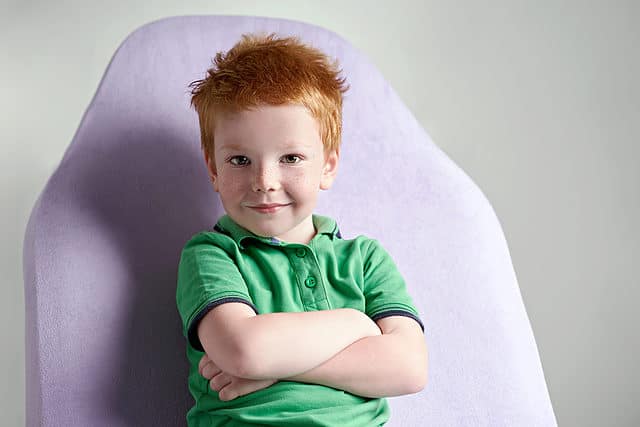 Cute red-haired freckled little boy wearing a green polo t-shirt sitting on a couch in a doctor's office waiting for a medical examination