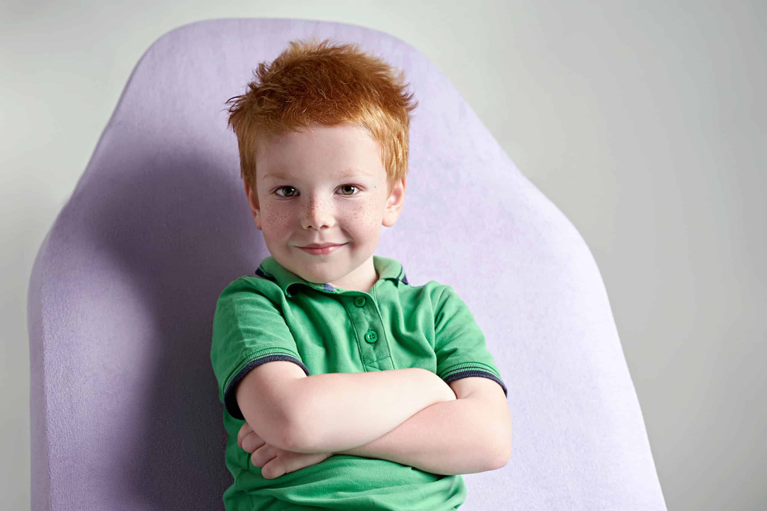 Cute red-haired freckled little boy wearing a green polo t-shirt sitting on a couch in a doctor's office waiting for a medical examination