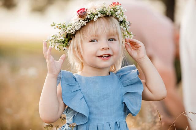 little girl is wearing a flower wreath on her head in a field on summer sunny day. baby in a blue dress.Portrait of adorable little child outdoors. happy holiday childhood.