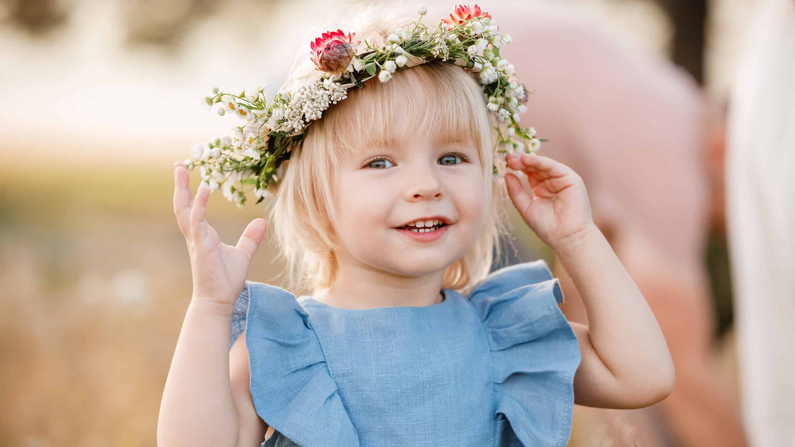 little girl is wearing a flower wreath on her head in a field on summer sunny day. baby in a blue dress.Portrait of adorable little child outdoors. happy holiday childhood.