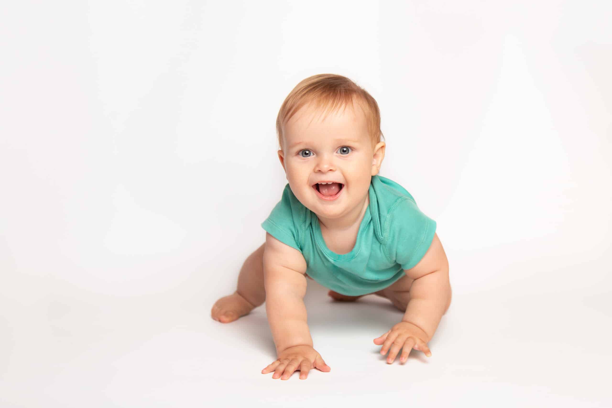 Cute small caucasian toddler baby boy child crawl on white studio floor. Smiling little infant kid wearing a green t-shirt explore world. Childcare and upbringing concept.