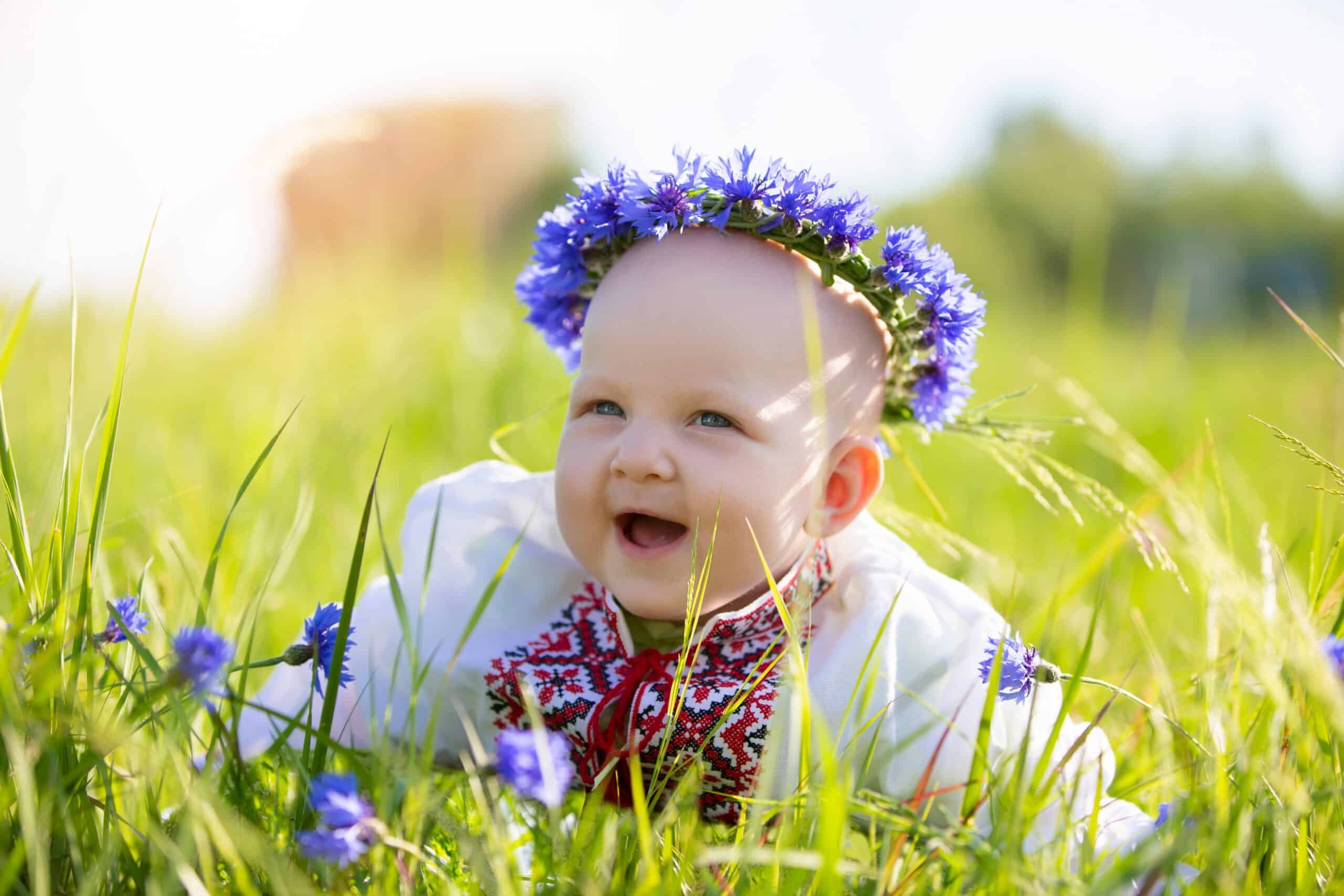 A happy little girl in national Belarusian or Ukrainian clothes lies on the grass and laughs. Beautiful Slavic child. A little baby in an embroidered shirt up to a year old on a summer meadow.