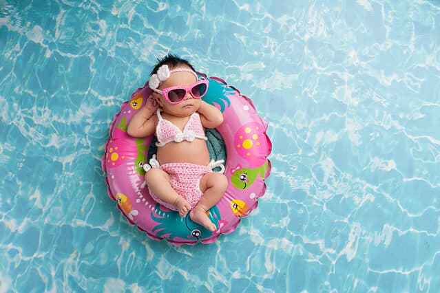 Nine day old newborn baby girl sleeping on a tiny inflatable swim ring. She is wearing a crocheted pink and white bikini and pink sunglasses.