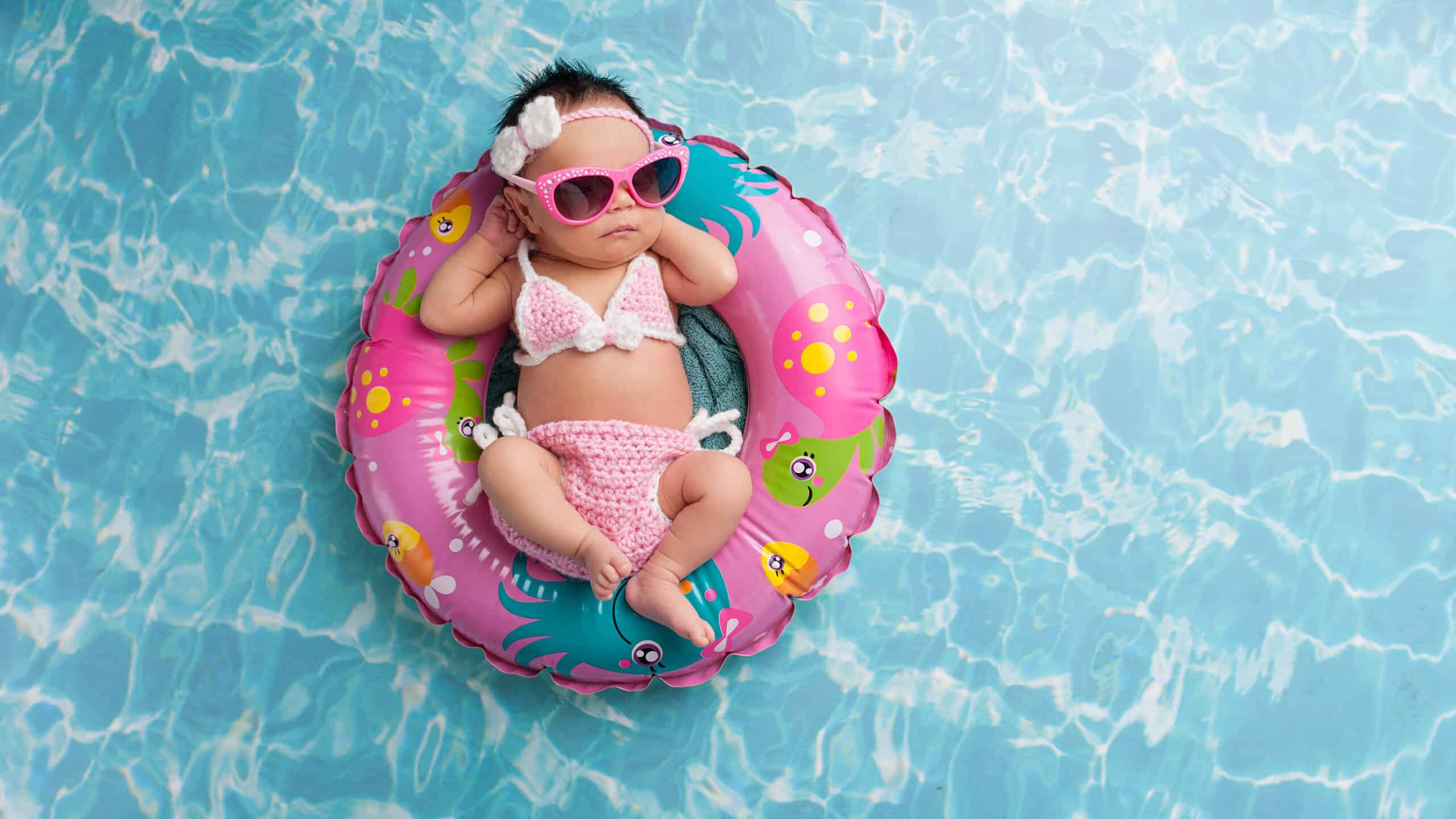 Nine day old newborn baby girl sleeping on a tiny inflatable swim ring. She is wearing a crocheted pink and white bikini and pink sunglasses.