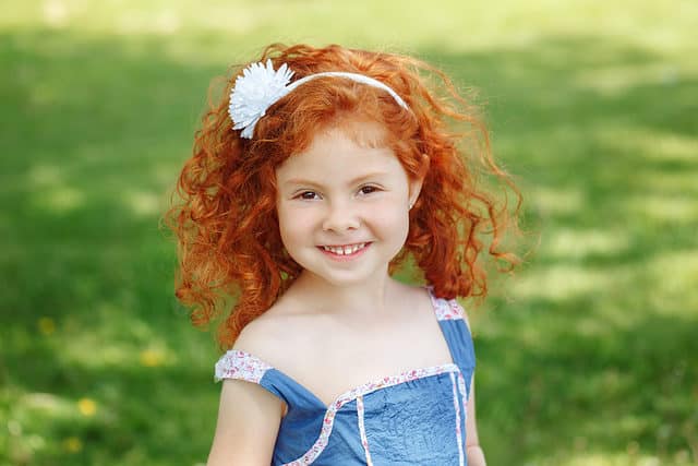 Closeup portrait of cute adorable little red-haired Caucasian girl child in a blue dress standing in a field meadow park outside looking into the camera, happy lifestyle childhood concept