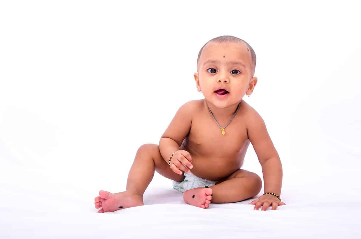 Happy looking nine months old infant Indian baby in a diaper, sitting isolated on a white background.